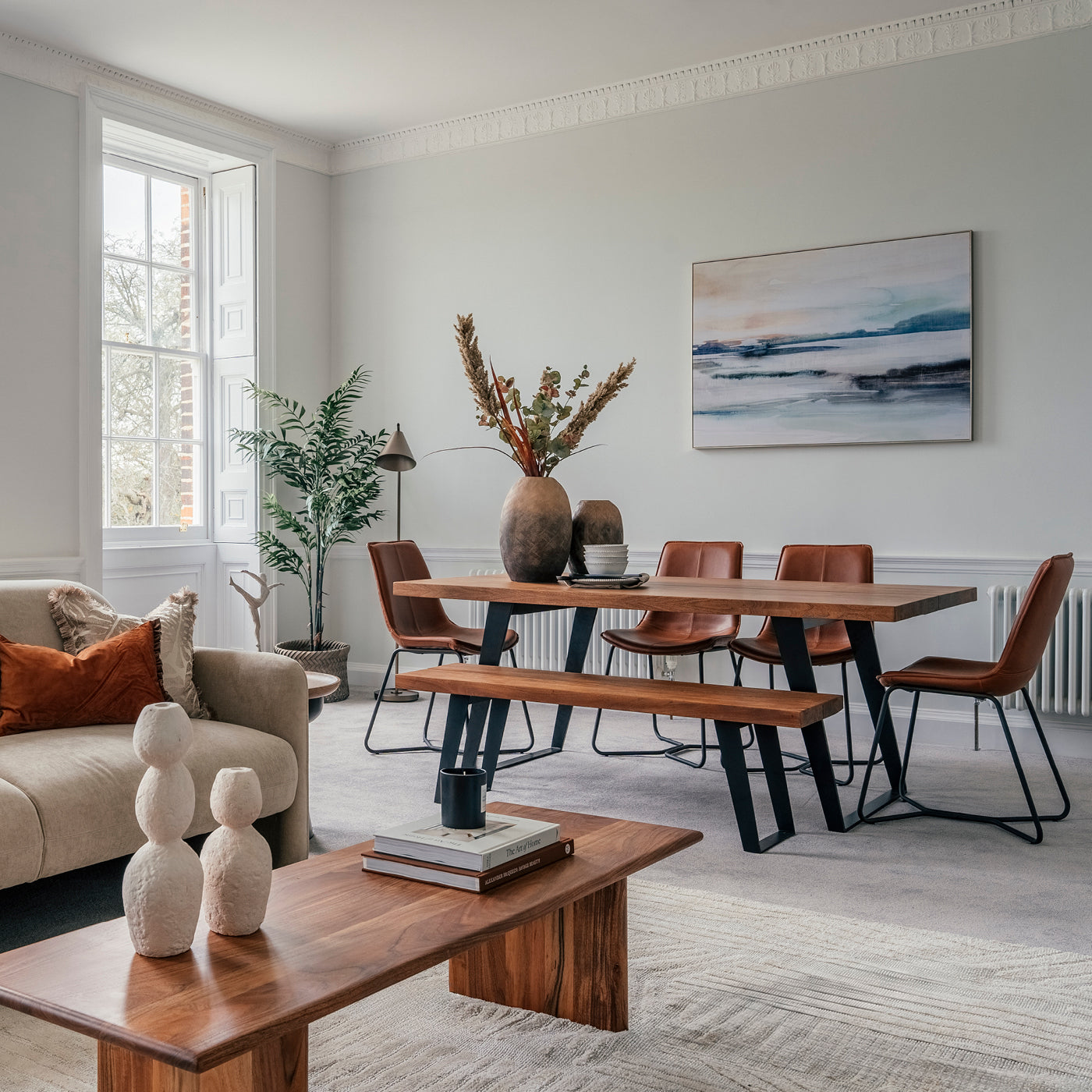 Contemporary dining and living space featuring a wooden dining table with leather chairs, a cozy beige sofa, and minimalist decor with earthy tones and abstract artwork.