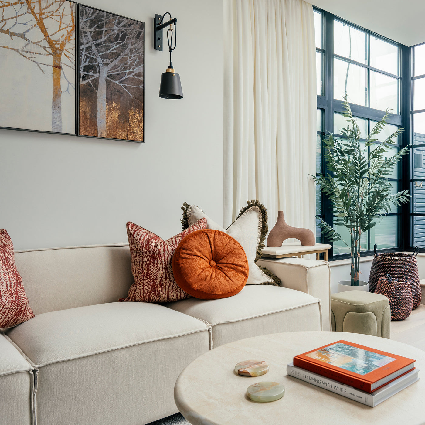 Modern living room with a cream sofa, colourful pillows, round coffee table with books, and large black-framed windows letting in natural light.