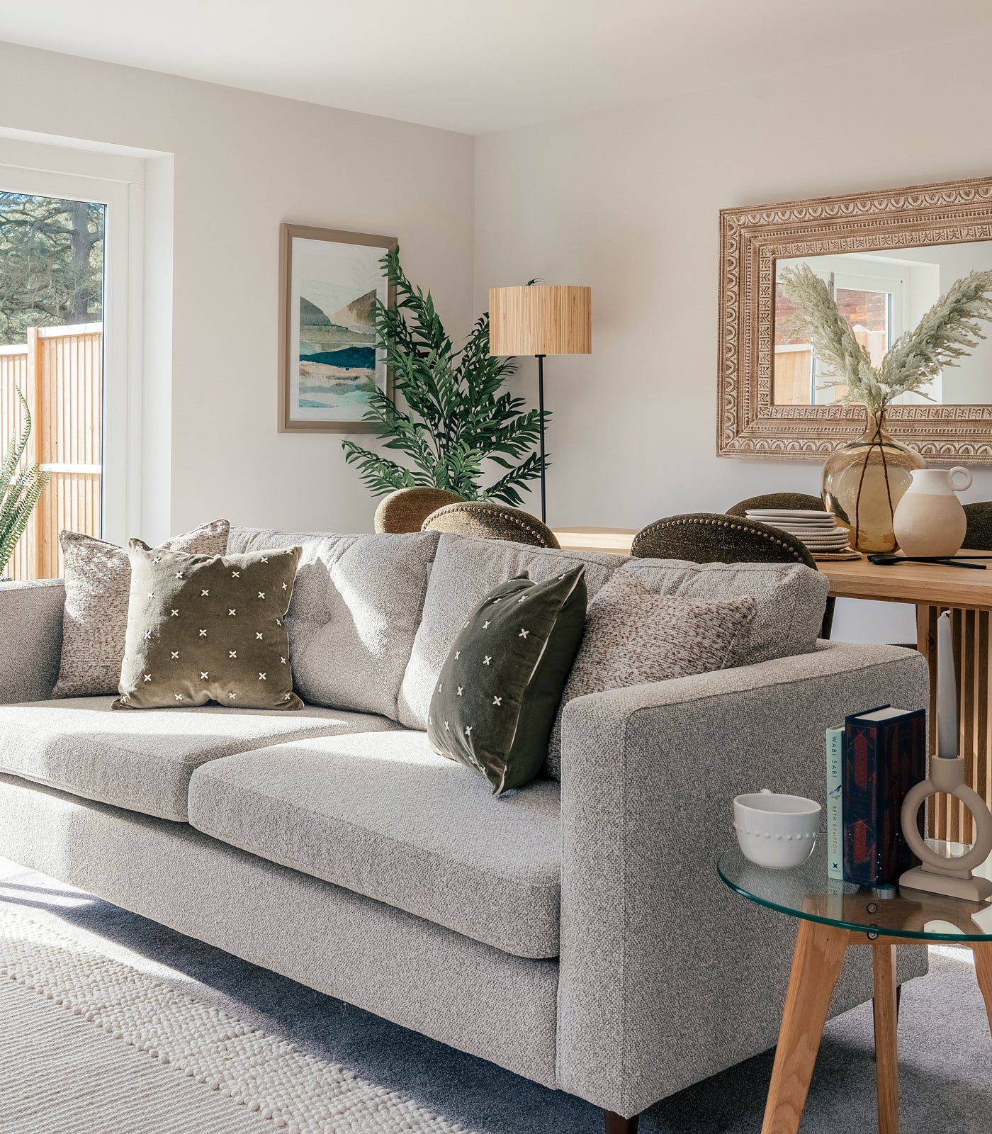 A cozy and well-lit living room with a light grey textured sofa adorned with green and patterned throw pillows. Behind the sofa, a wooden dining table with green upholstered chairs is complemented by a large decorative mirror and a glass vase with pampas grass. A framed landscape artwork and a tall floor lamp stand beside a leafy plant in the corner. A small round glass side table holds a ceramic cup, books, and a sculptural candle holder, adding a personal touch to the inviting space.