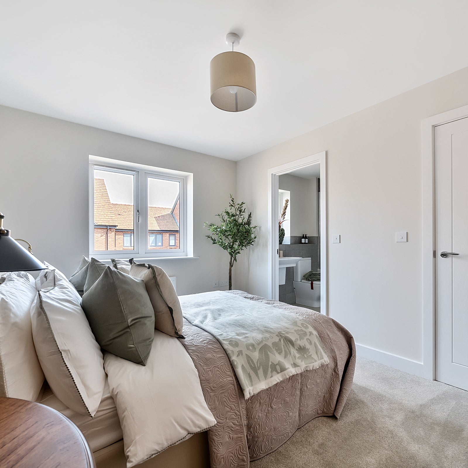 Bright and airy bedroom with a double bed, soft neutral tones, green accent pillows, and an ensuite bathroom visible through an open door.