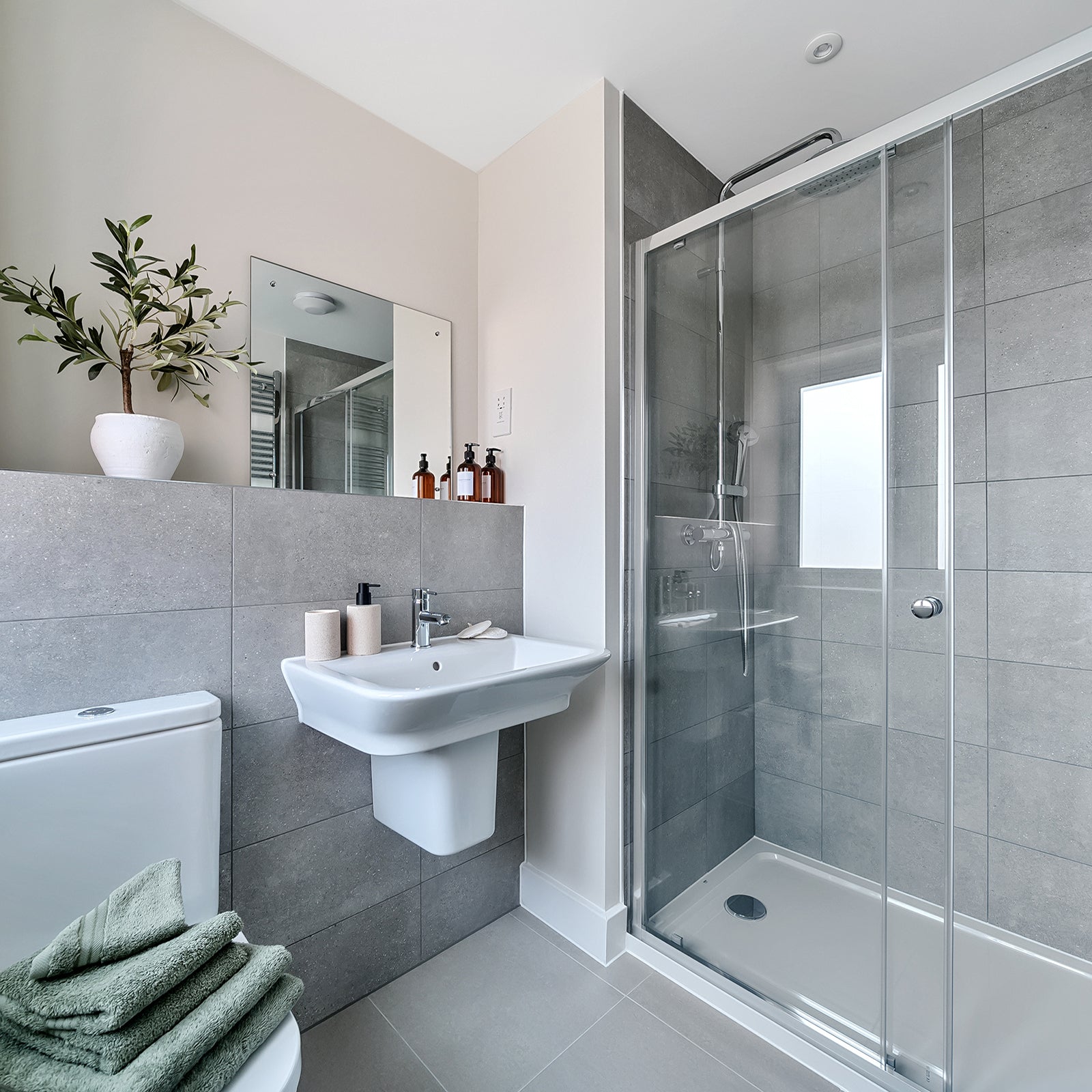 Contemporary bathroom featuring a glass-enclosed shower, wall-mounted basin, grey tiling, and minimalist décor including a potted plant and folded green towels.