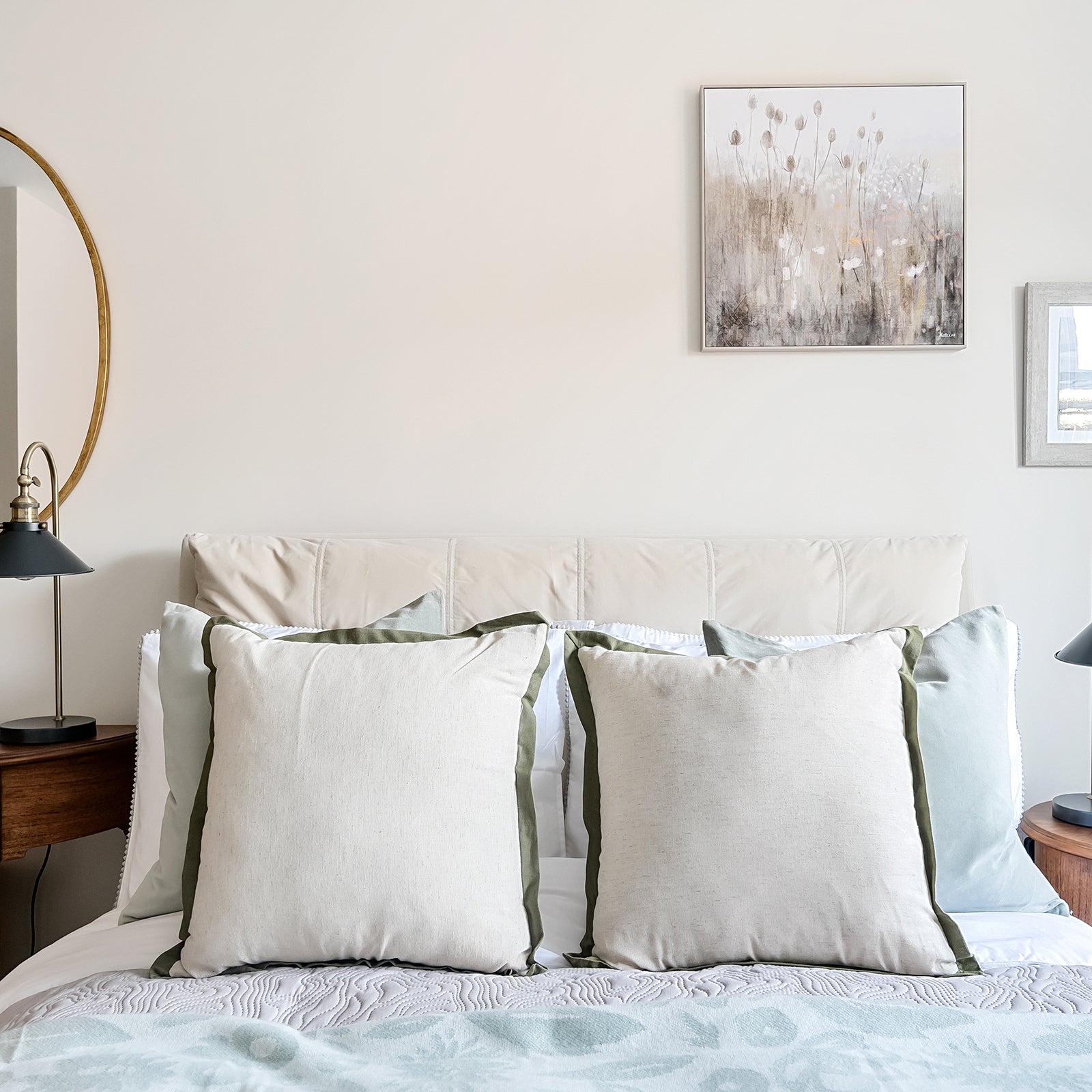 Close-up of a neatly made bed with large cream and sage green cushions, a quilted throw, and matching bedside tables with black reading lamps, set against a neutral wall with framed artwork.