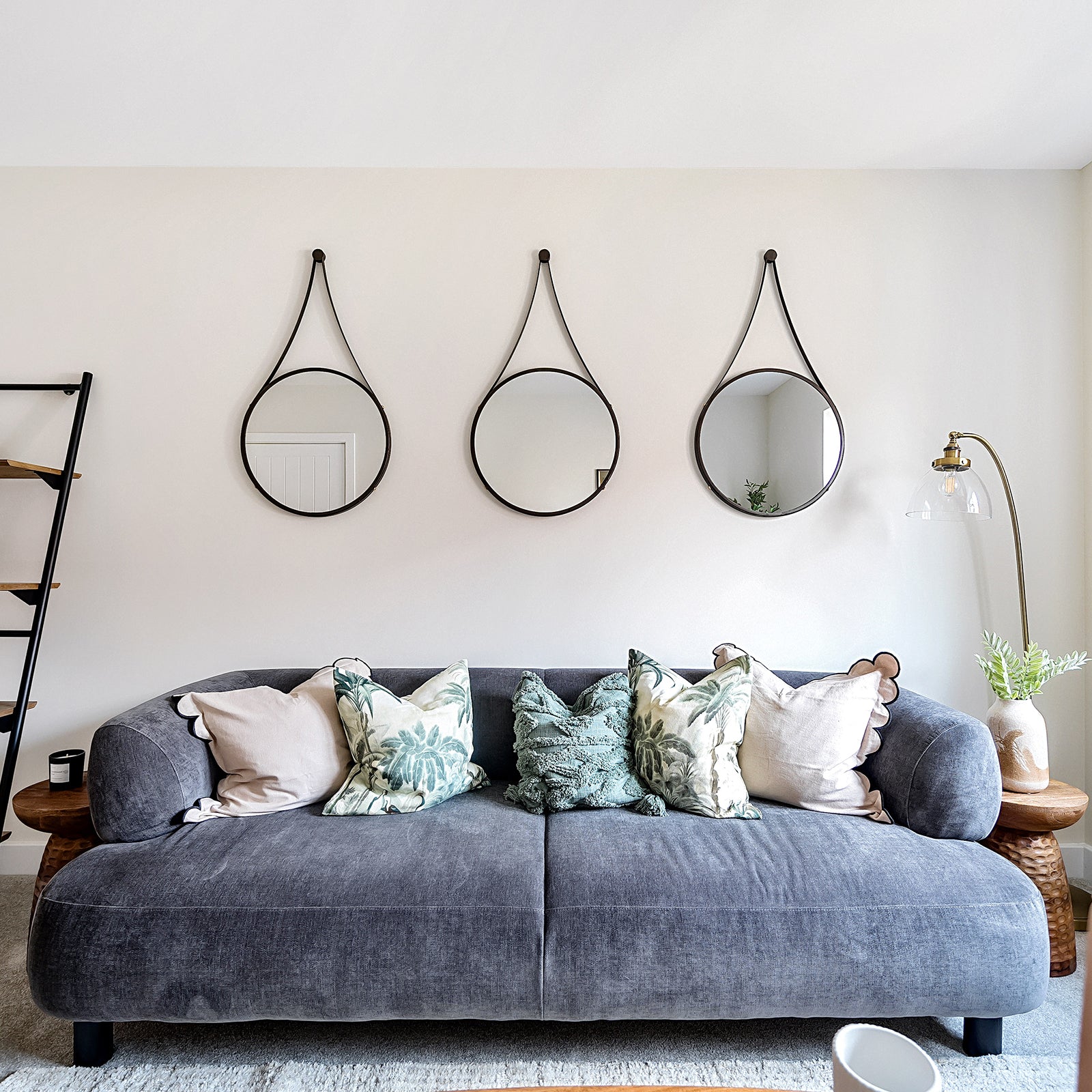 Stylish living room featuring a deep grey velvet sofa with assorted botanical and neutral cushions, set against a cream wall adorned with three round mirrors in black leather-look straps, flanked by wooden side tables and a brass floor lamp.