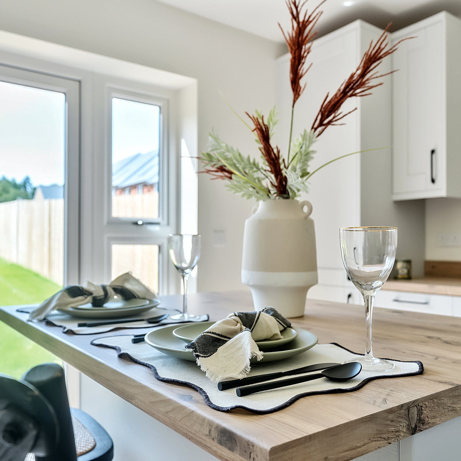 Close-up of a modern kitchen breakfast bar set for two, featuring sage green plates, black cutlery, and fringed napkins on cream placemats, with a large ceramic vase of dried and faux foliage as a centrepiece, and natural light streaming through French doors overlooking a garden.