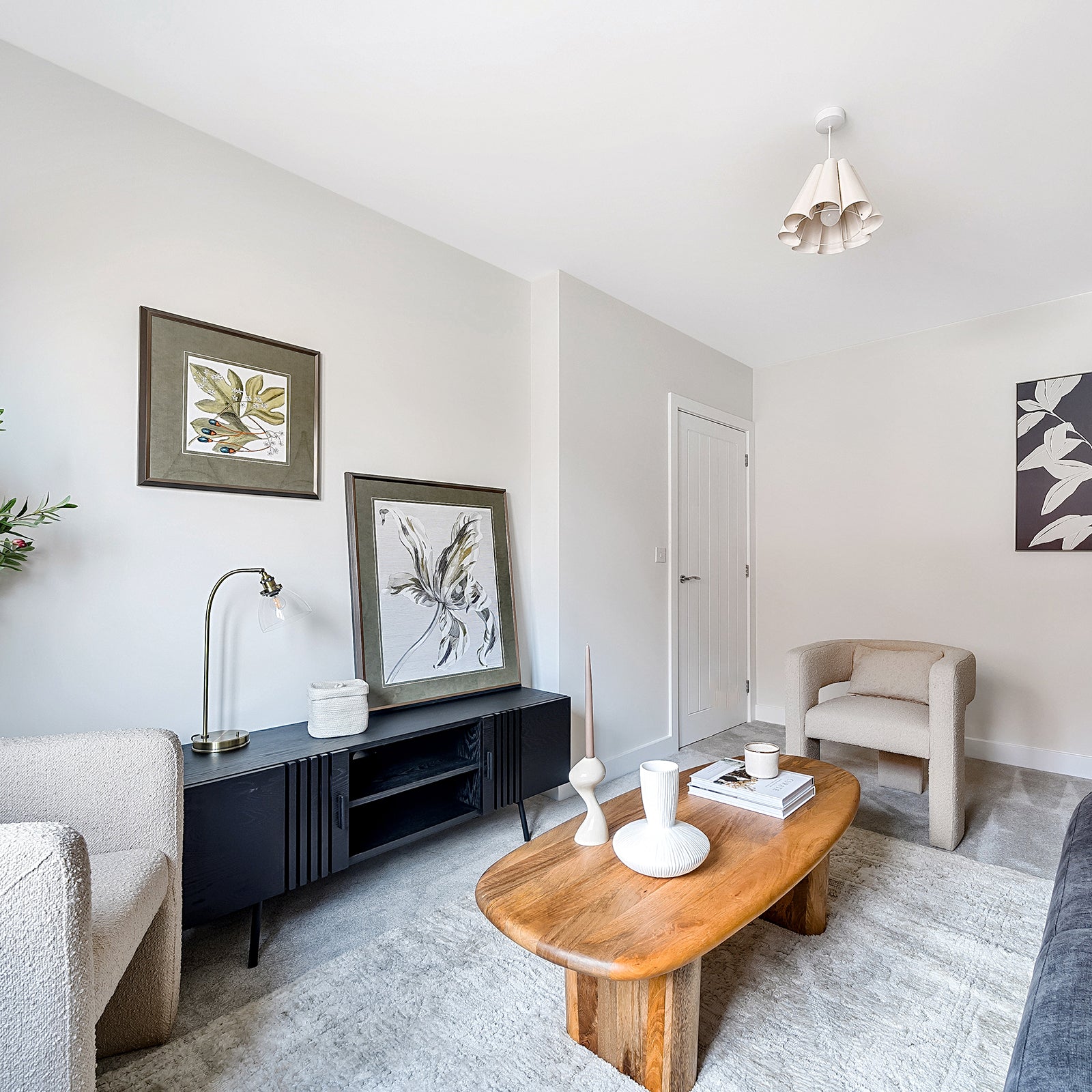 Modern sitting room with neutral tones, featuring a textured beige armchair, black sideboard with framed botanical artwork, a wooden coffee table styled with minimalist white vases and books, all set against light walls and a soft grey rug.