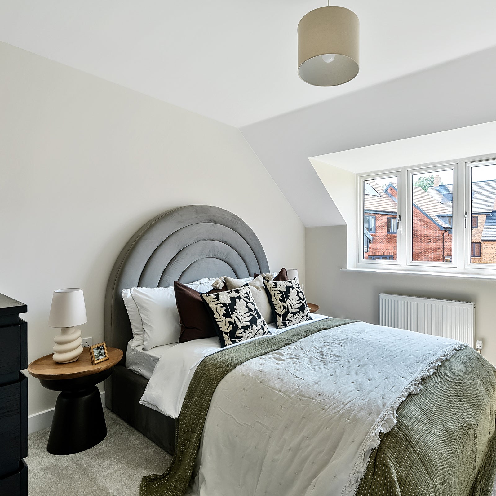 Contemporary bedroom with a large arched grey velvet headboard, layered bedding in white and olive green, decorative cushions in bold leaf prints, and a stylish bedside table with a sculptural lamp, set under a sloped ceiling with a view through a window to red-brick houses outside.