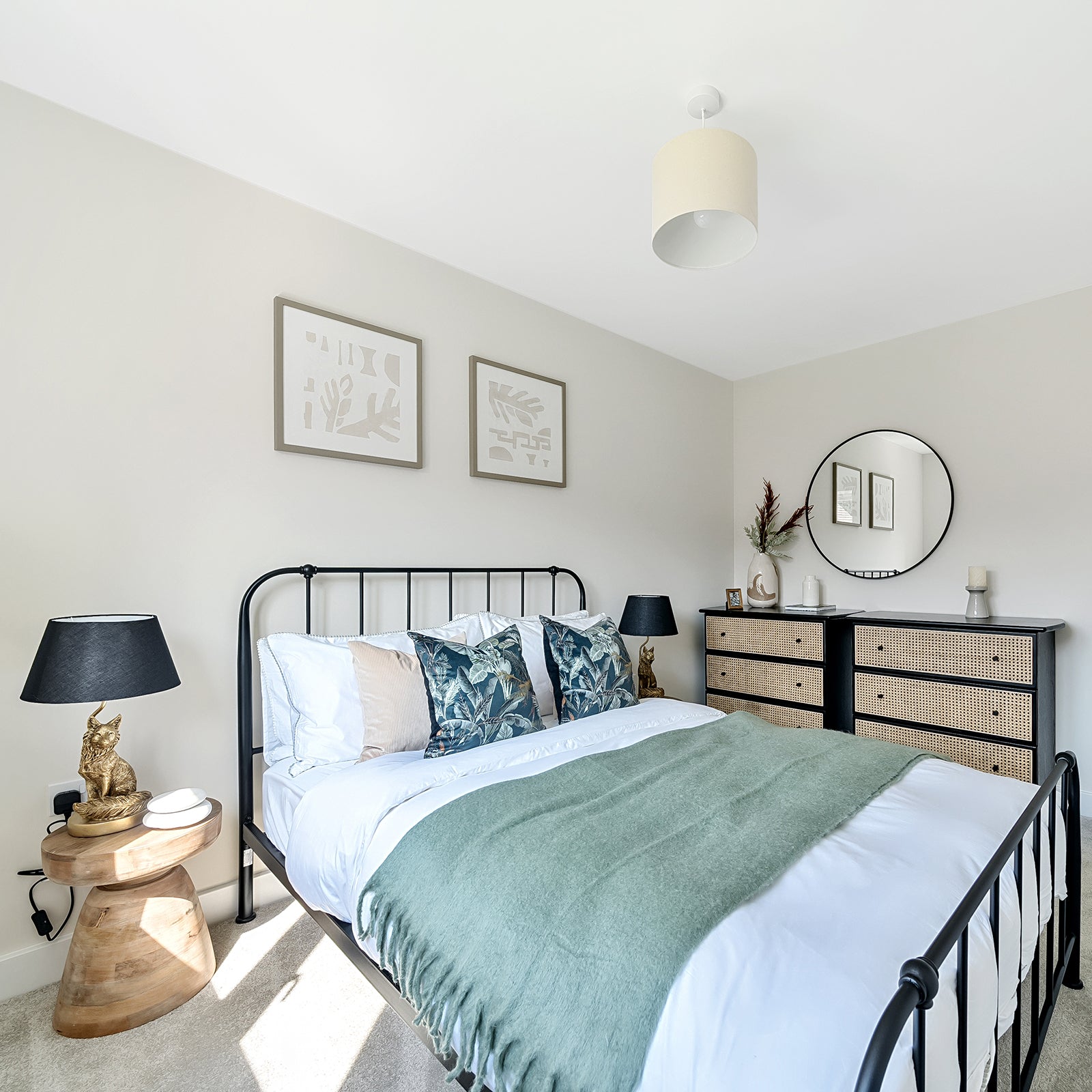Stylish bedroom featuring a black metal bed frame with white bedding and green accent throw, complemented by tropical print and neutral cushions; natural wood and woven furnishings include a bedside table with a gold rabbit lamp and black shade, and a chest of drawers with a circular mirror above.