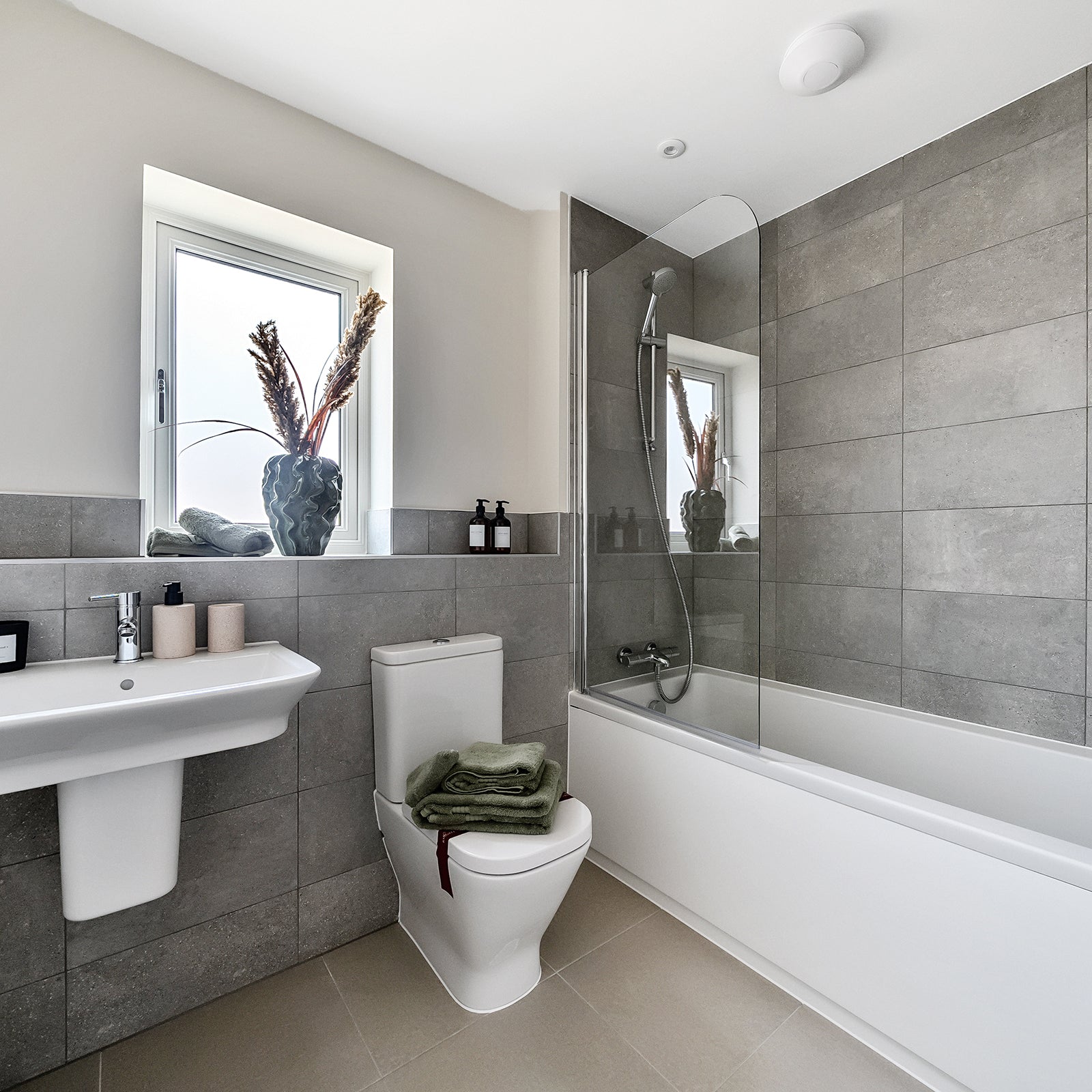 Contemporary bathroom decorated in neutral tones, featuring a wall-mounted white basin, a toilet with folded green towels placed on top, and a bath with a sleek glass shower screen; grey tiled walls and a window with a decorative vase of dried grasses add a stylish finishing touch.
