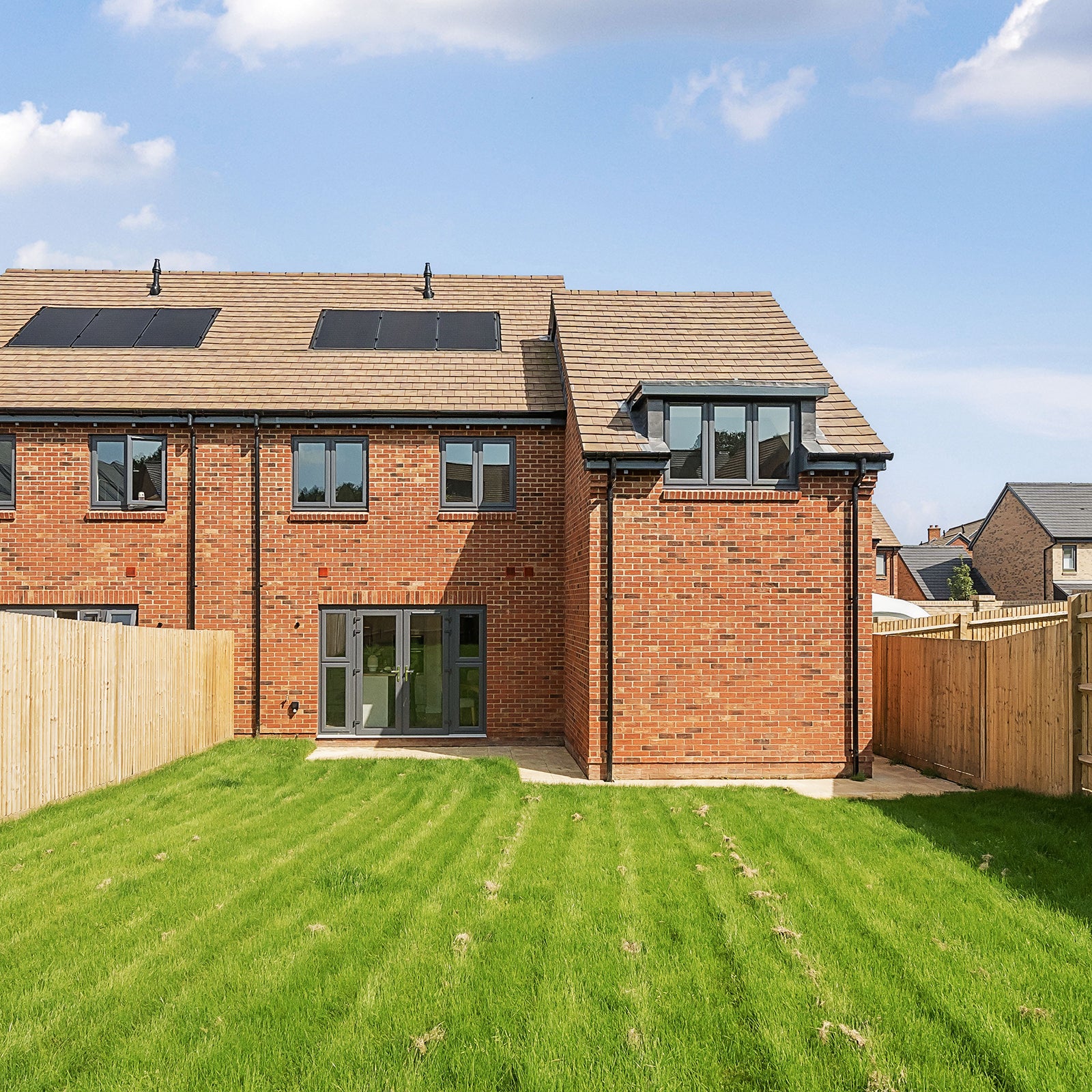 Rear view of a modern red-brick semi-detached house with a pitched tiled roof, grey-framed windows, and solar panels; featuring a spacious enclosed garden with a neatly mown lawn and a patio area outside the double doors.