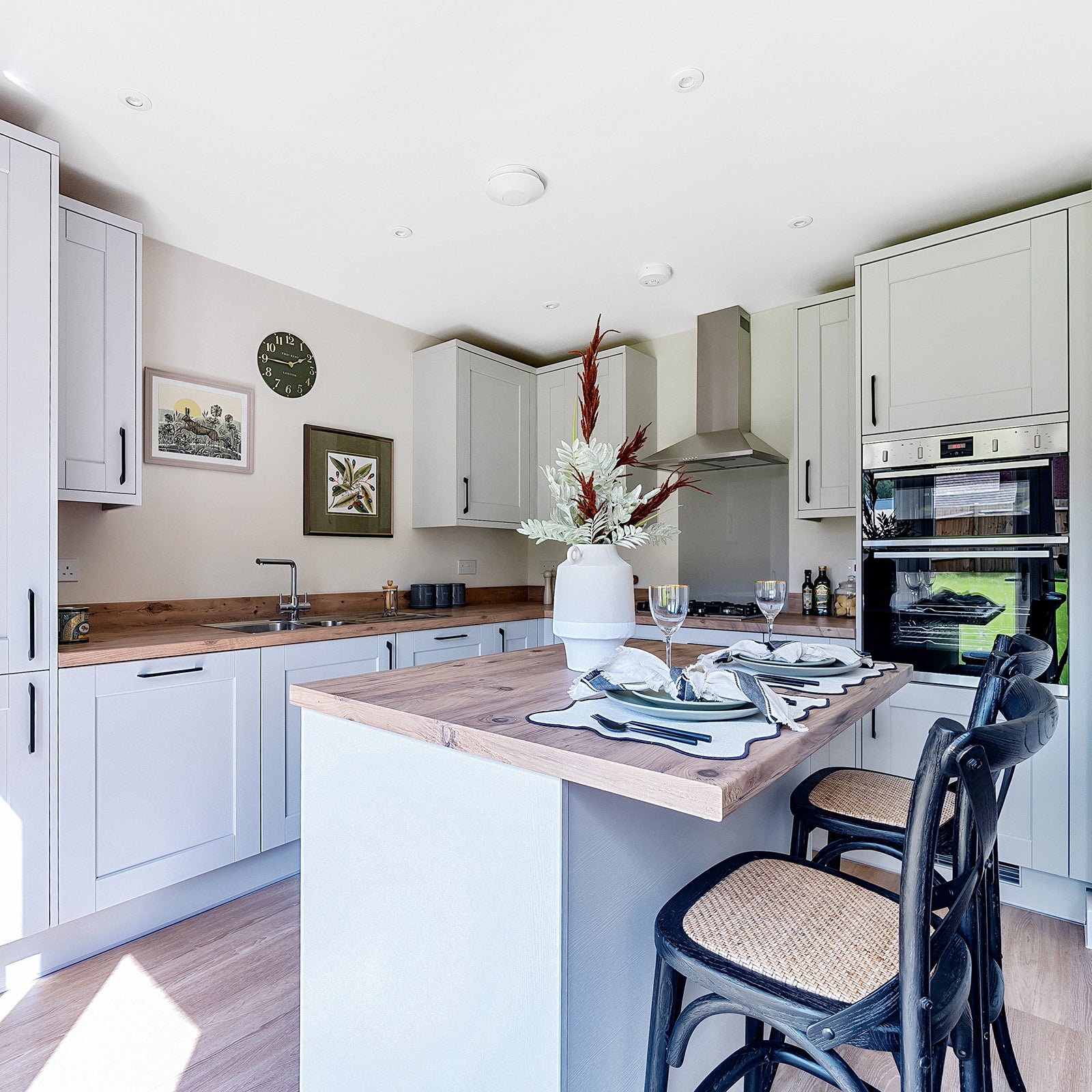 Modern kitchen with light grey shaker-style cabinets, wood-effect countertops, and integrated stainless steel appliances including a double oven; featuring a central island with a wooden worktop, black bar stools with woven seats, and a decorative vase with foliage as a centrepiece.
