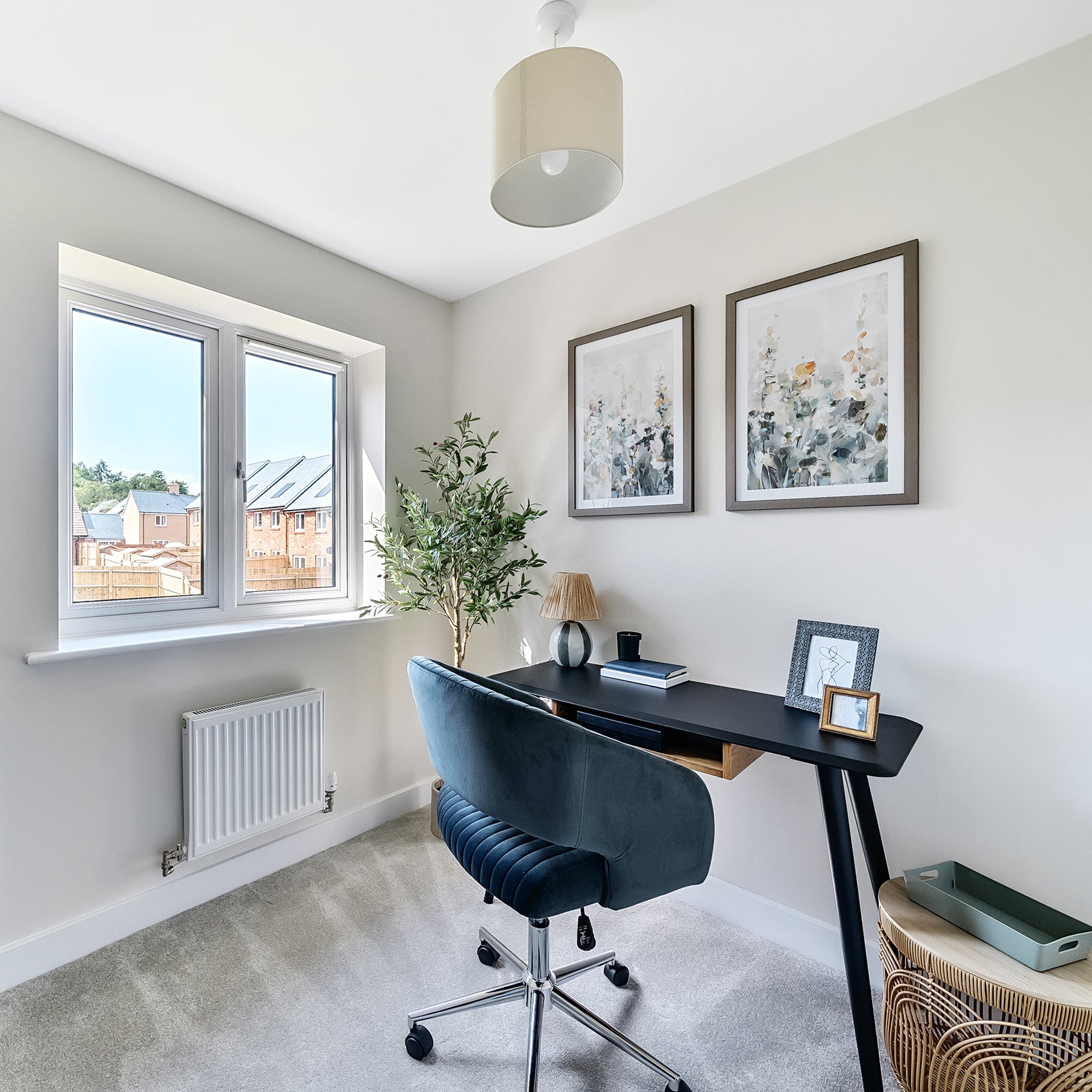 Contemporary home office with a large window allowing natural light to fill the space, featuring a sleek black desk, a blue velvet swivel chair, neutral carpet, and wall-mounted abstract artwork above the desk; accessorised with a potted plant, small table lamp, and decorative photo frames.