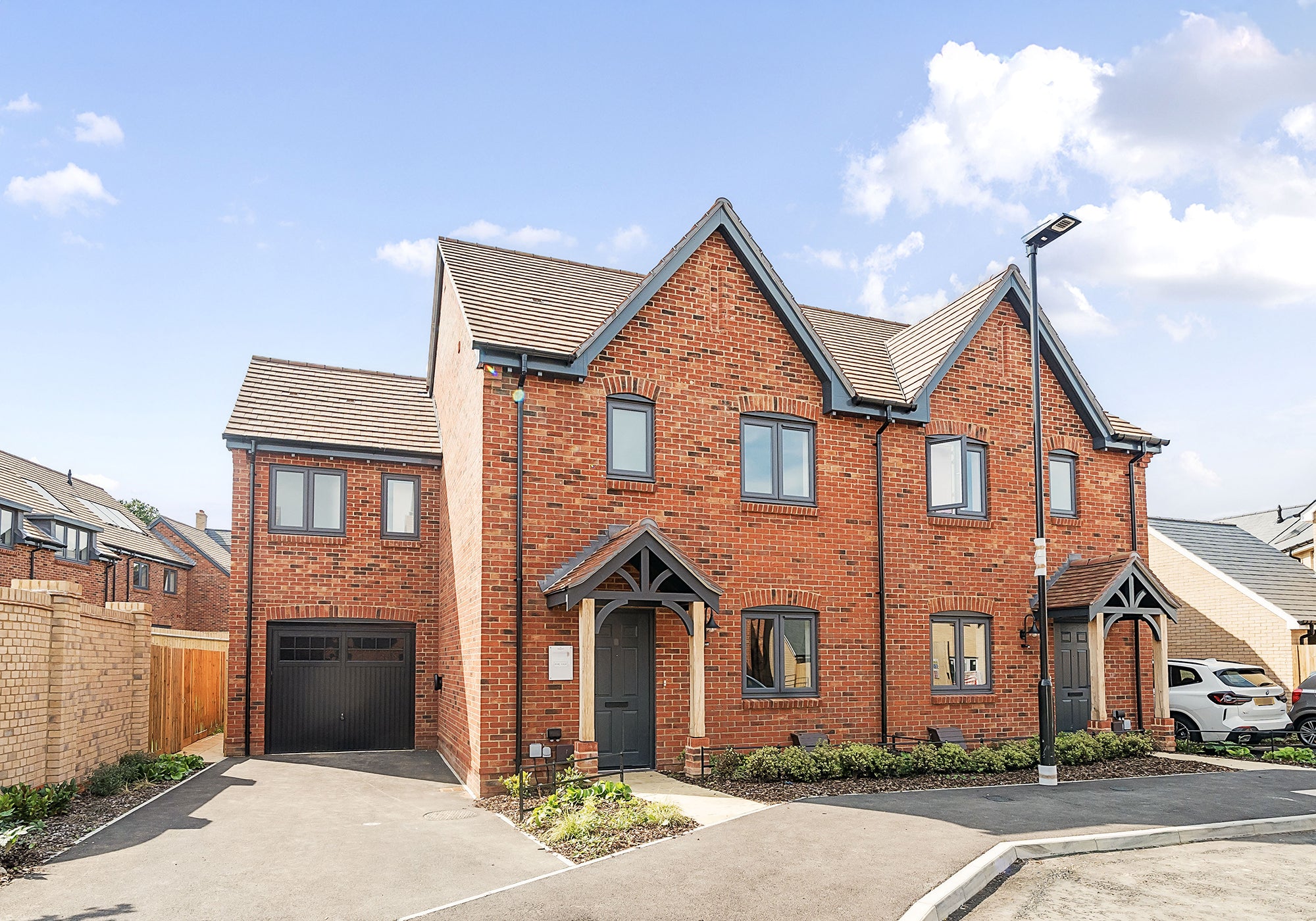 Front view of a modern semi-detached red-brick house with pitched roofs, grey framed windows and doors, a single garage to the left, and a neatly maintained driveway and front garden, all under a clear blue sky.