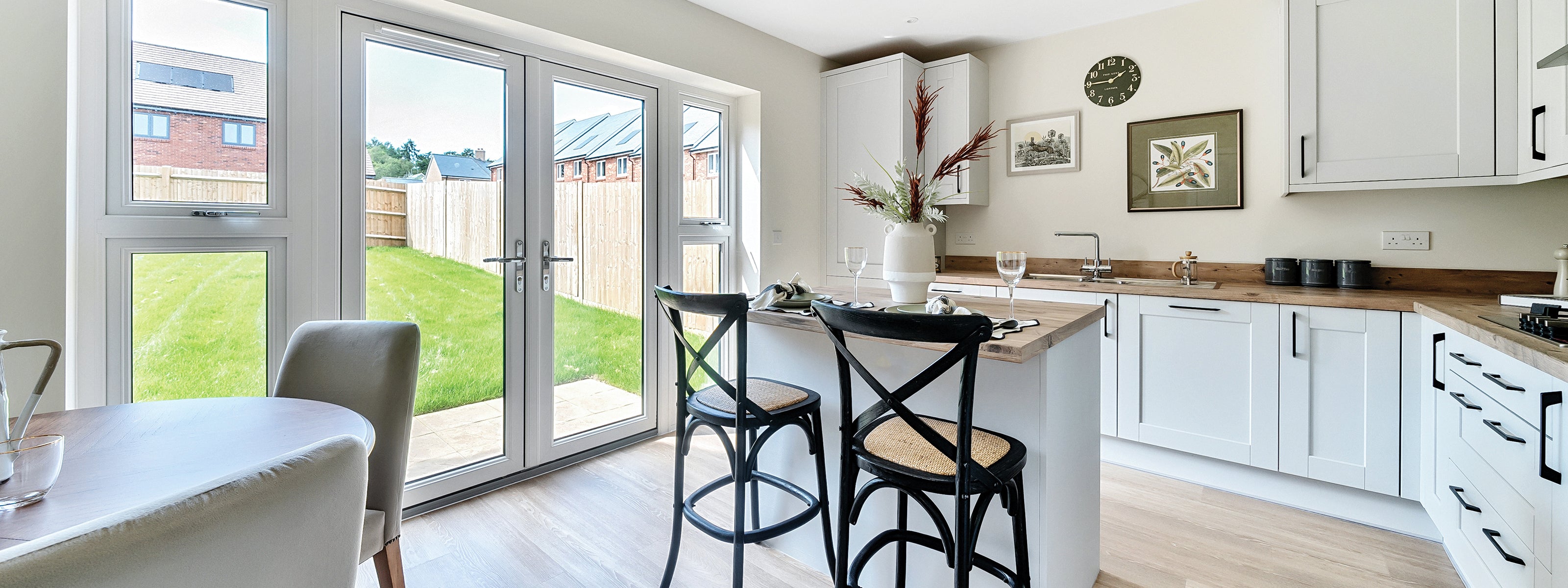 Bright and modern kitchen interior featuring a central island with two bar stools, shaker-style white cabinetry, wood countertops, wall-mounted artwork and a clock, with large French doors and windows opening to a private grassy garden.