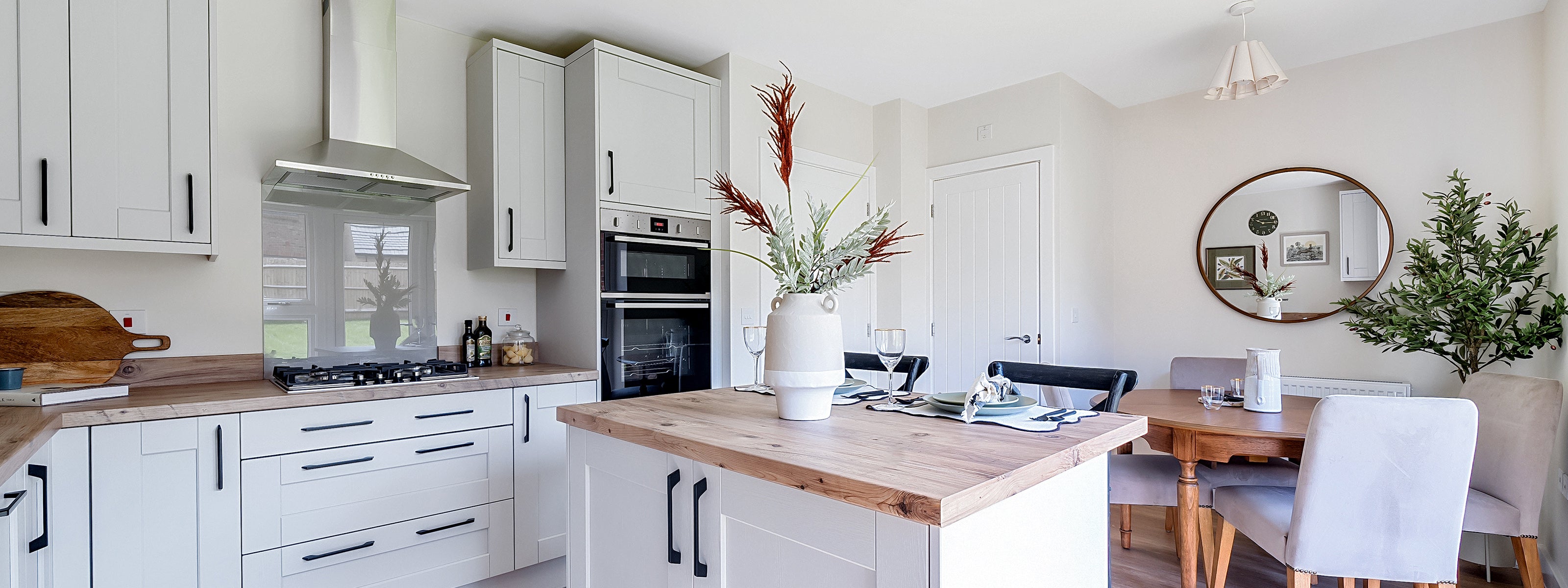 Contemporary kitchen and dining area with white shaker-style cabinets, wooden countertops, built-in double oven, and gas hob, centred around an island set for dining with a vase of flowers; adjacent is a wooden dining table with upholstered chairs, a round wall mirror, and a tall indoor plant adding a touch of greenery.