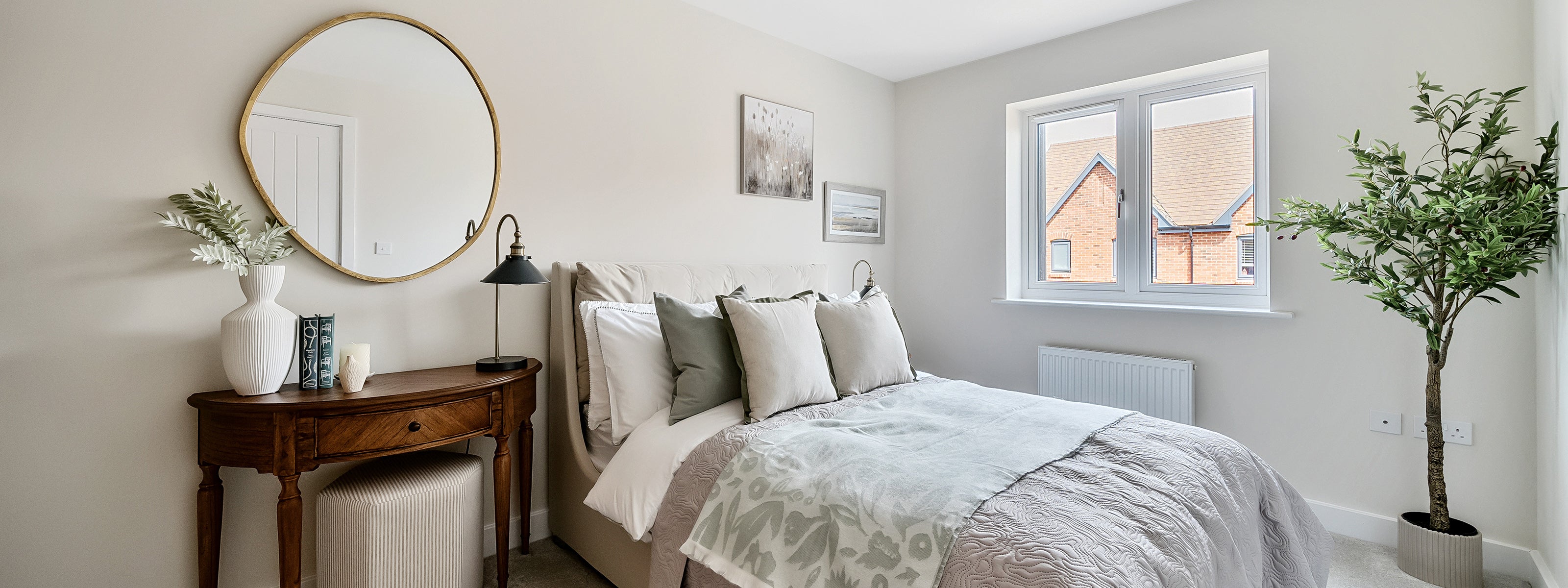 Bright and serene bedroom featuring a neatly made bed with neutral-toned linens and multiple cushions, a wooden vanity table with a round wall mirror above, decorative accents including a white vase and books, and a potted plant adding a natural touch near a large window with views of neighbouring brick houses.
