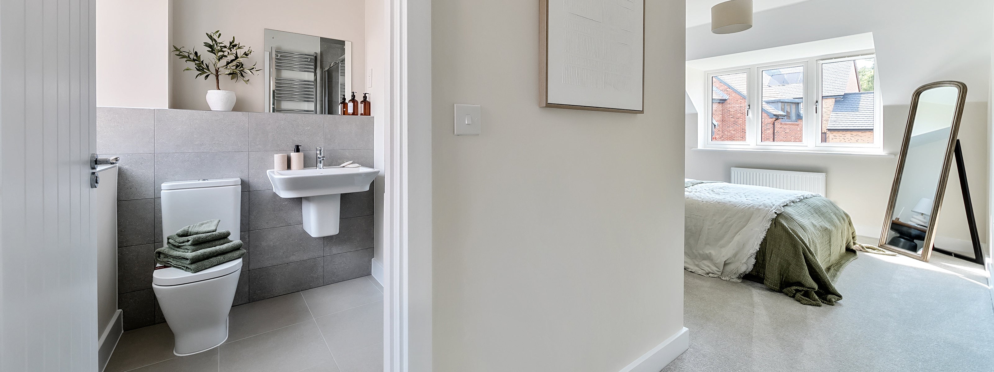 View of a modern, minimalist bathroom and adjoining bedroom; the bathroom features grey tiled walls, a white toilet with stacked green towels, a pedestal sink with toiletries, and a mirror above, while the bedroom has a bed with green and white bedding, a large angled mirror, and a window letting in natural light onto the soft carpeted floor.