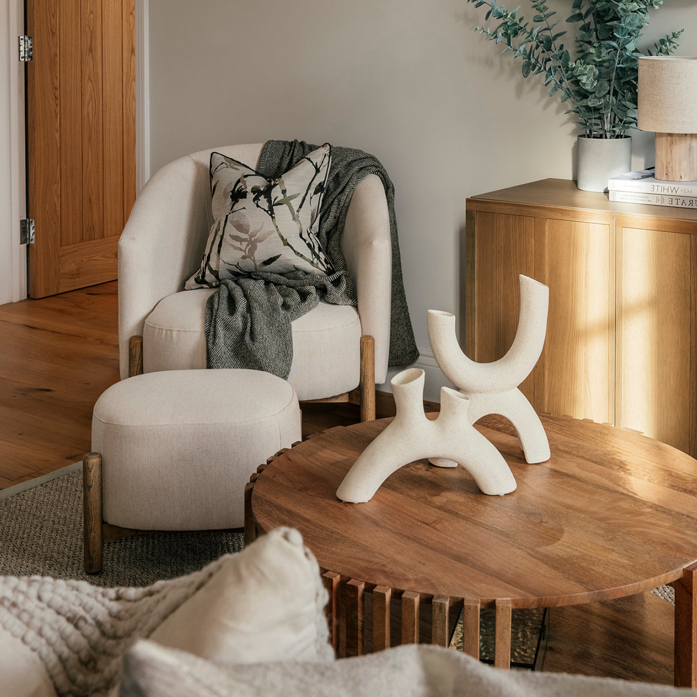 A cozy reading nook featuring a cream upholstered armchair with matching ottoman, adorned with a botanical-patterned pillow and a draped grey throw. The space includes a round wooden coffee table with sculptural ceramic decor, a wooden sideboard topped with books, a lamp, and a potted plant, all set against a neutral-toned wall and warm wooden flooring.
