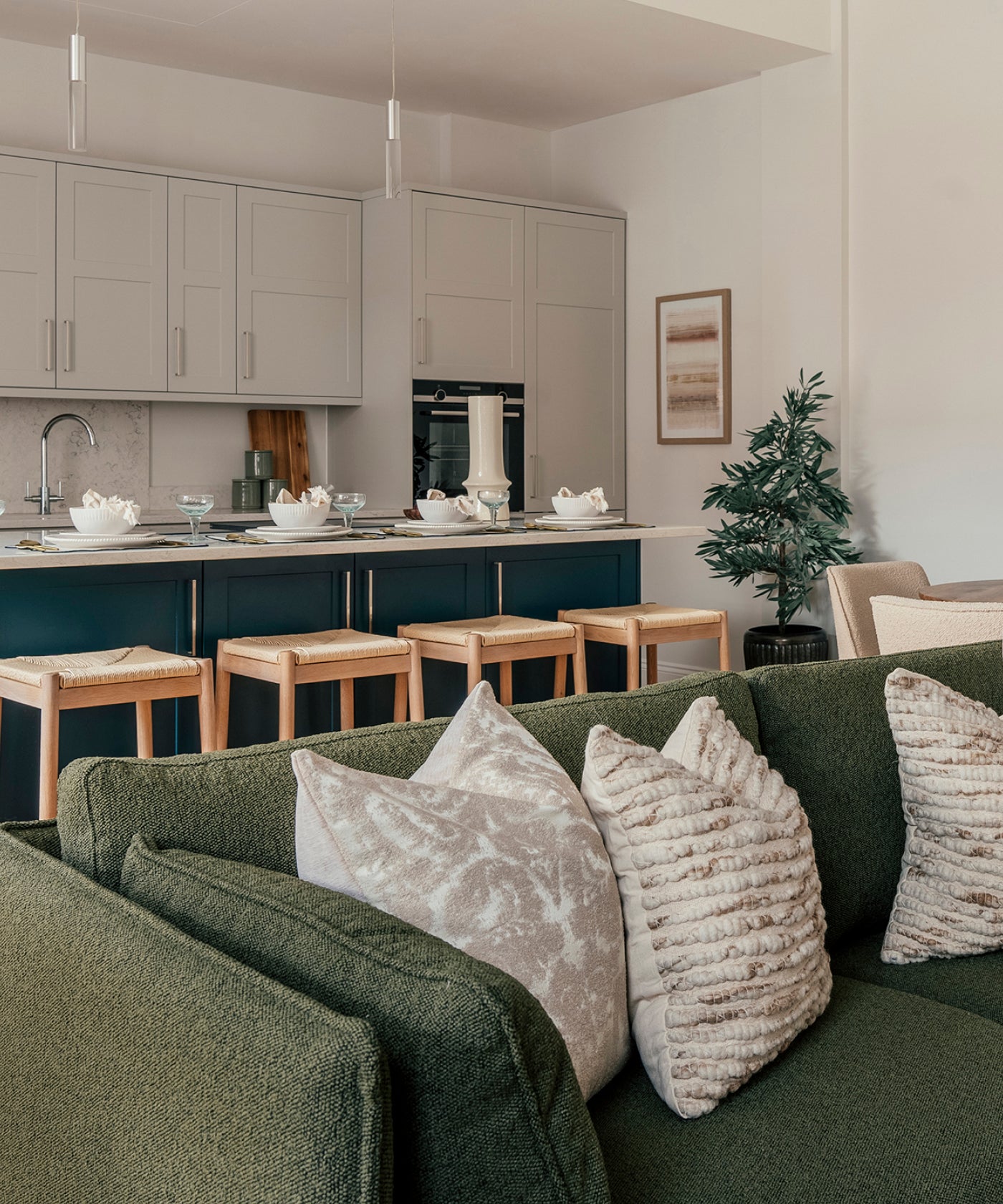 A modern open-plan kitchen and living area featuring a deep green textured sofa with plush neutral-toned cushions in the foreground. Behind the sofa, a stylish kitchen includes a blue island with wooden bar stools, beige cabinetry, a marble backsplash, and a breakfast setup with elegant dishware. A potted plant and framed artwork add warmth and decoration to the neutral-toned space.