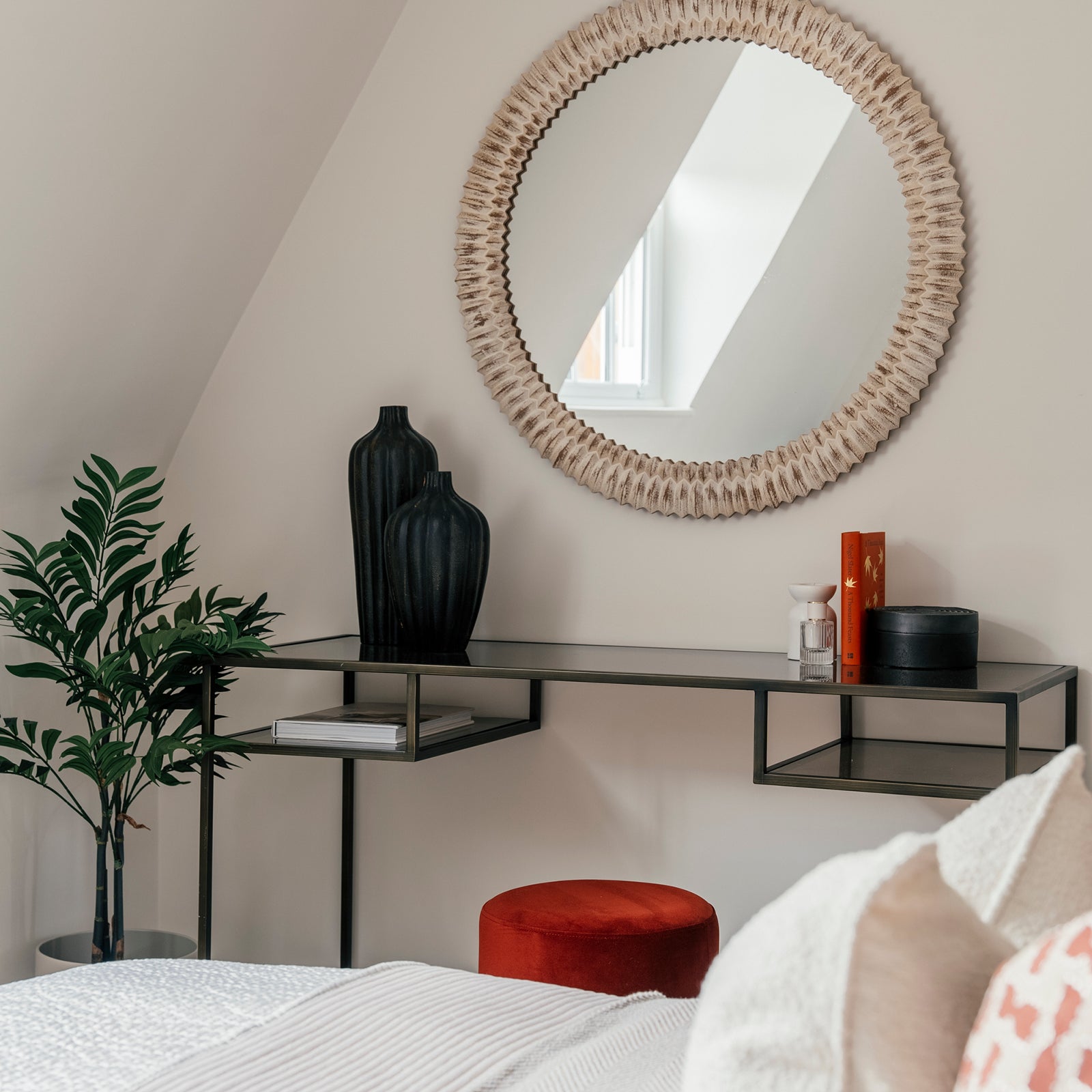 A stylish bedroom corner with a black metal console table holding two black vases, orange books, a diffuser, and a round black box. Above it hangs a large round mirror with a textured beige frame. A red velvet stool sits below the table, next to a green potted plant. The bed in the foreground features white and cream linens with a hint of coral.