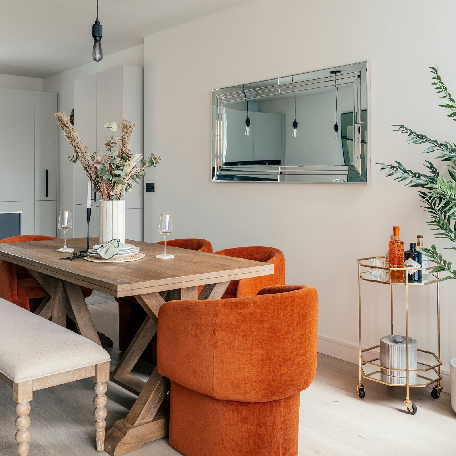 A stylish dining area featuring a rustic wooden table surrounded by plush burnt-orange chairs and a beige upholstered bench. A tall vase with dried florals serves as the centrepiece, while a large mirrored wall piece reflects the room. A gold bar cart with assorted bottles and a potted plant sit to the right, adding elegance to the neutral-toned space.