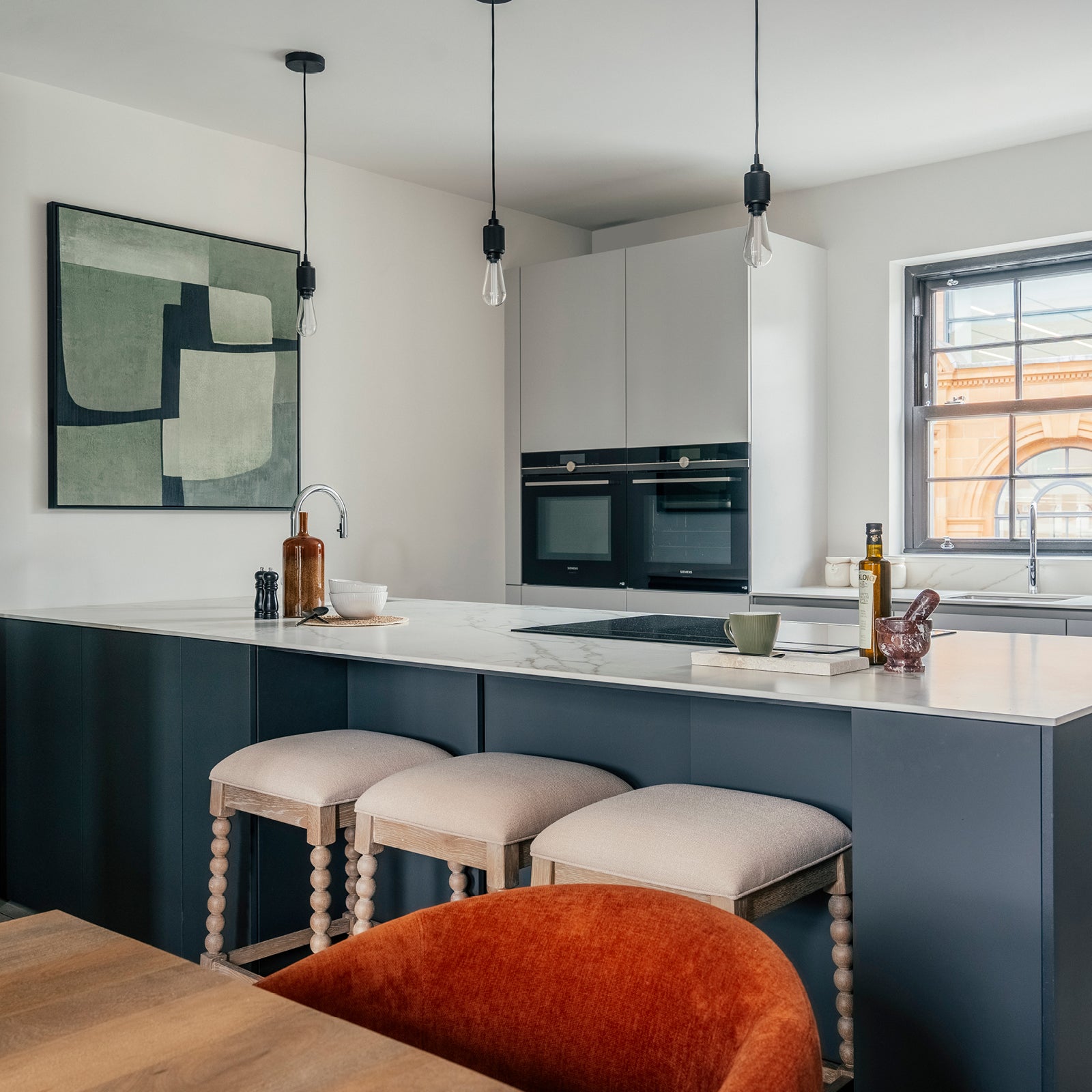 A modern kitchen with a white marble island, dark blue cabinetry, and three cushioned wooden bar stools. Pendant lights hang above, and built-in double ovens are set against a matte cabinet wall. A window adds natural light, with minimal decor and abstract art completing the space.