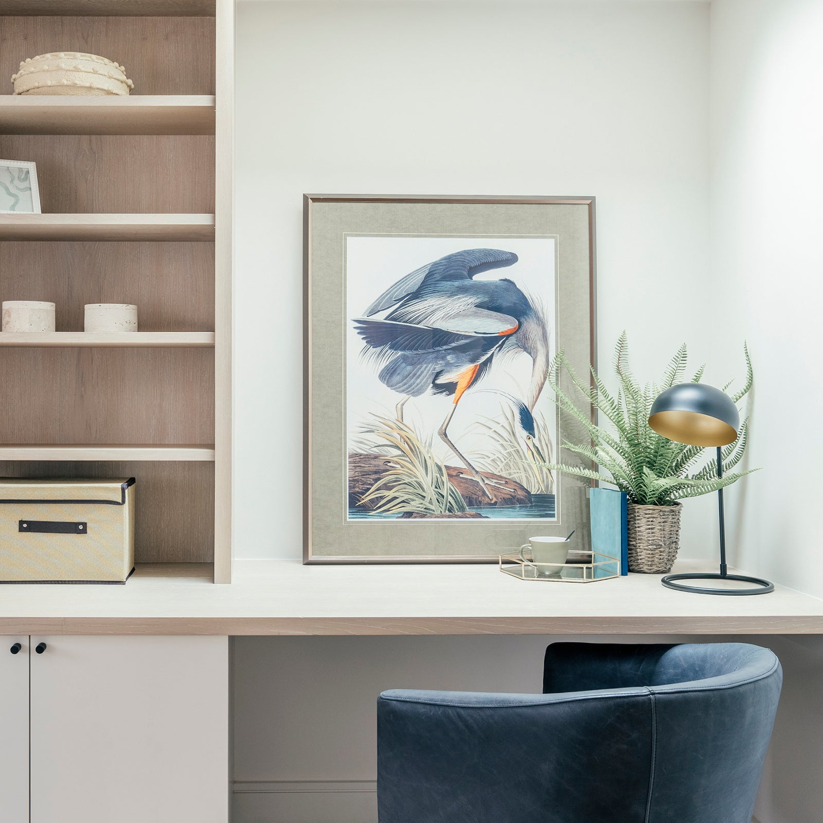 A compact home office nook featuring a wooden desk, open shelving, a framed bird print, and a black-and-gold desk lamp, with a blue leather chair and a potted fern adding colour and texture.