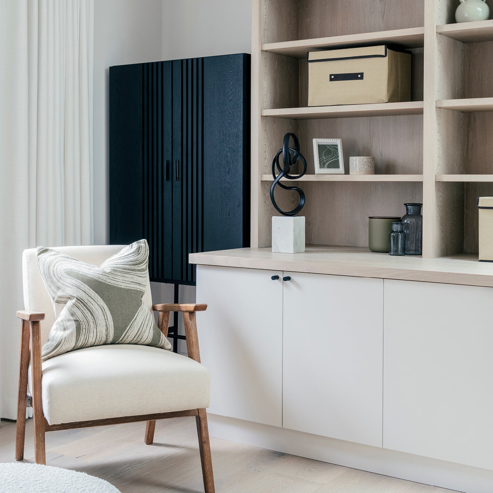 Minimalist home office corner featuring a cream armchair with a patterned cushion, light wood built-in shelving with decorative accents and storage boxes, and a bold black cabinet adding visual contrast.