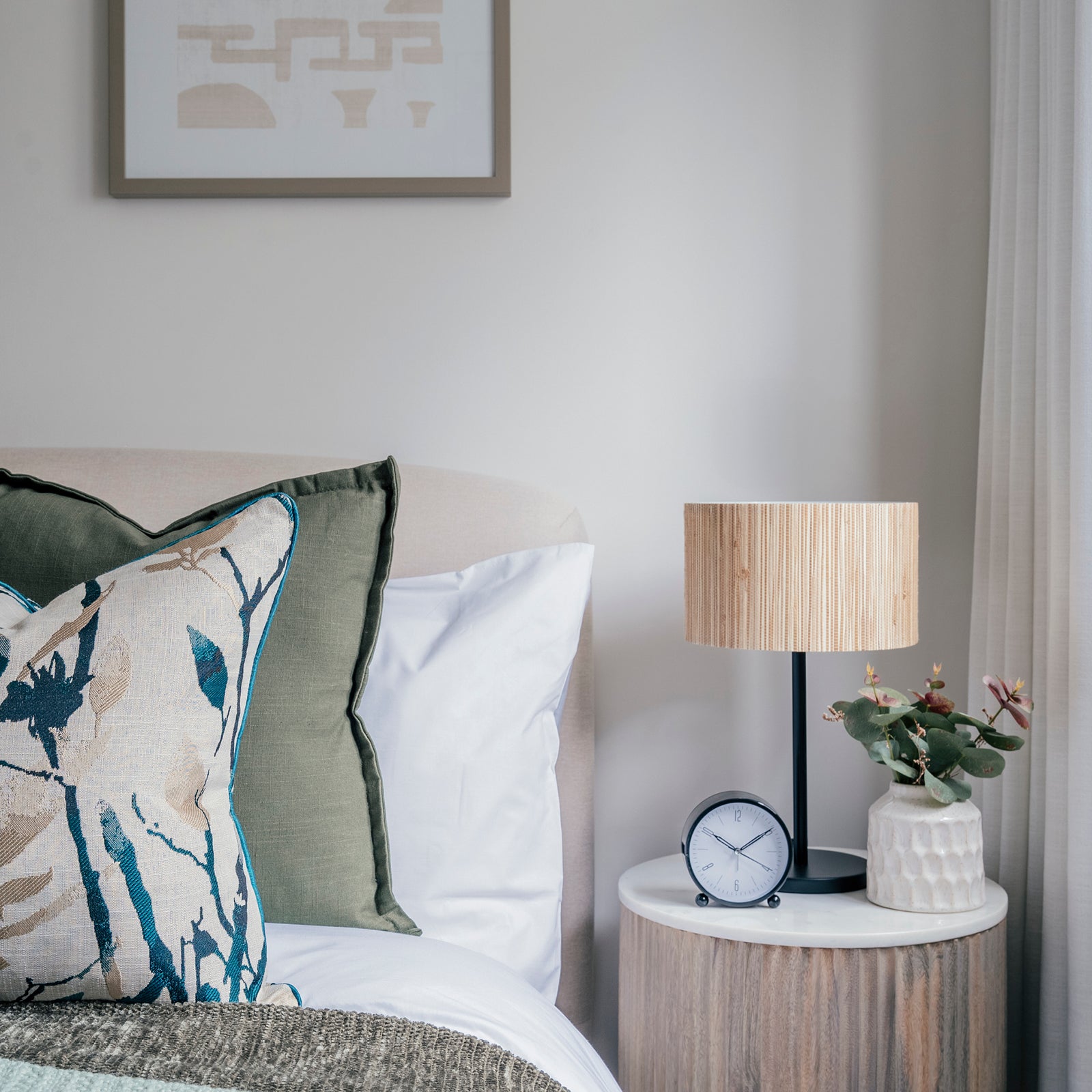Calm bedroom scene with a soft upholstered headboard, botanical accent pillows, and a round wooden bedside table topped with a modern table lamp, a small plant, and a classic black-rimmed clock.