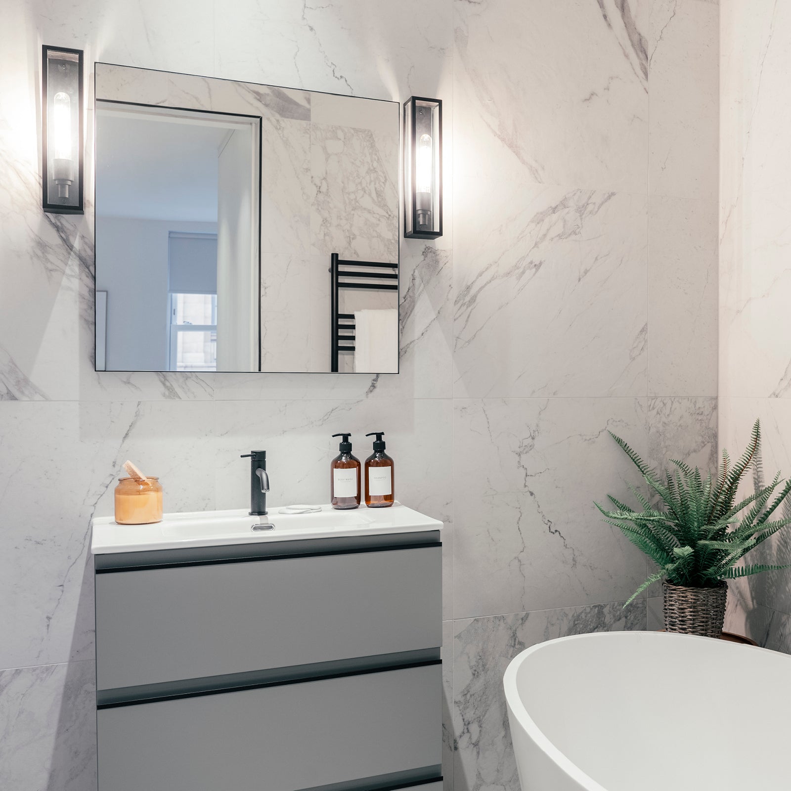 Modern bathroom with marble-look tiled walls, a rectangular mirror flanked by sleek black wall sconces, a minimalist grey vanity with a black faucet, amber soap dispensers, a white freestanding bathtub, and a potted fern adding a touch of greenery.