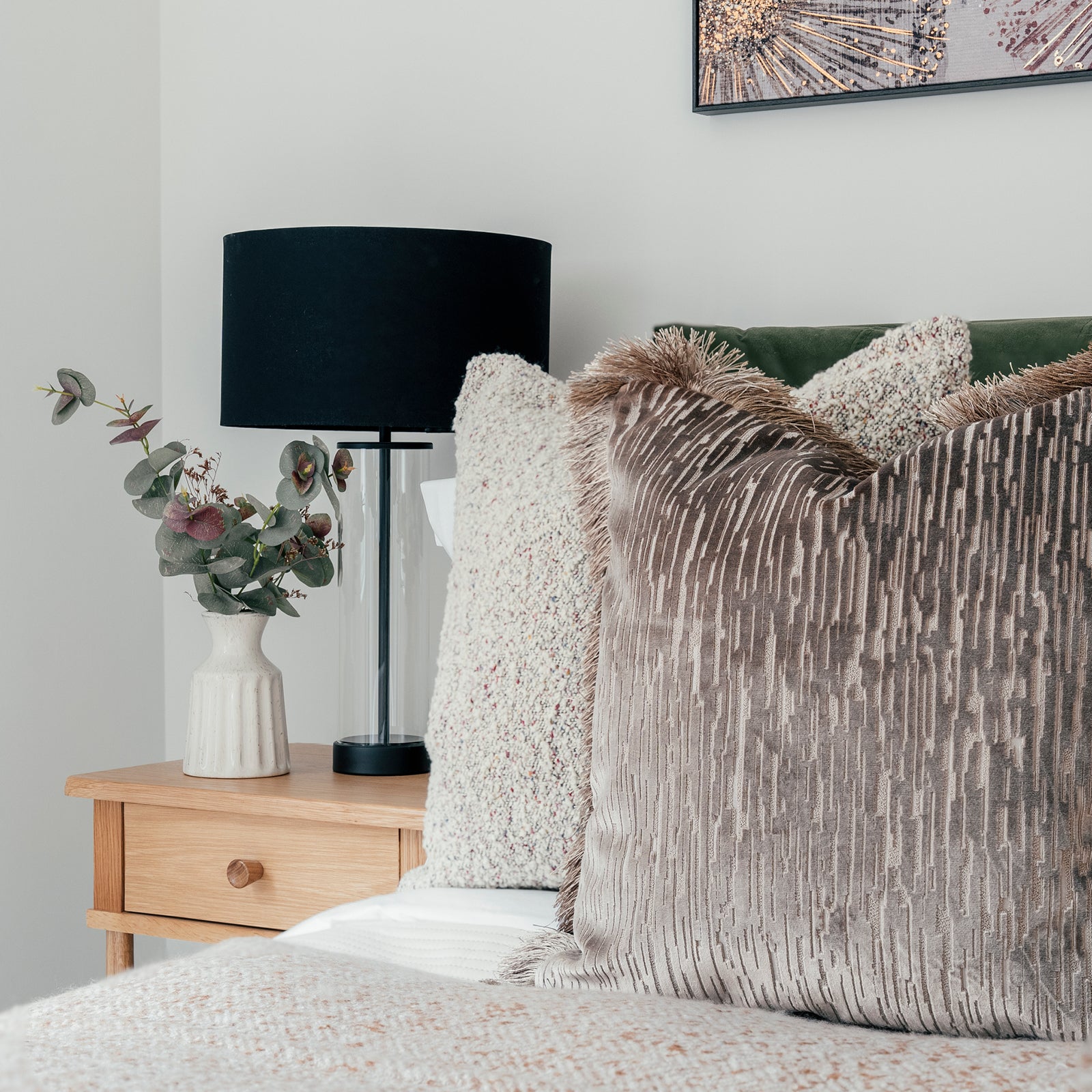 Close-up of a cozy bedroom corner featuring a wooden nightstand with a textured ceramic vase of eucalyptus branches, a black lampshade with a glass base, and a bed adorned with plush, textured throw pillows in neutral and metallic tones against a green velvet headboard.