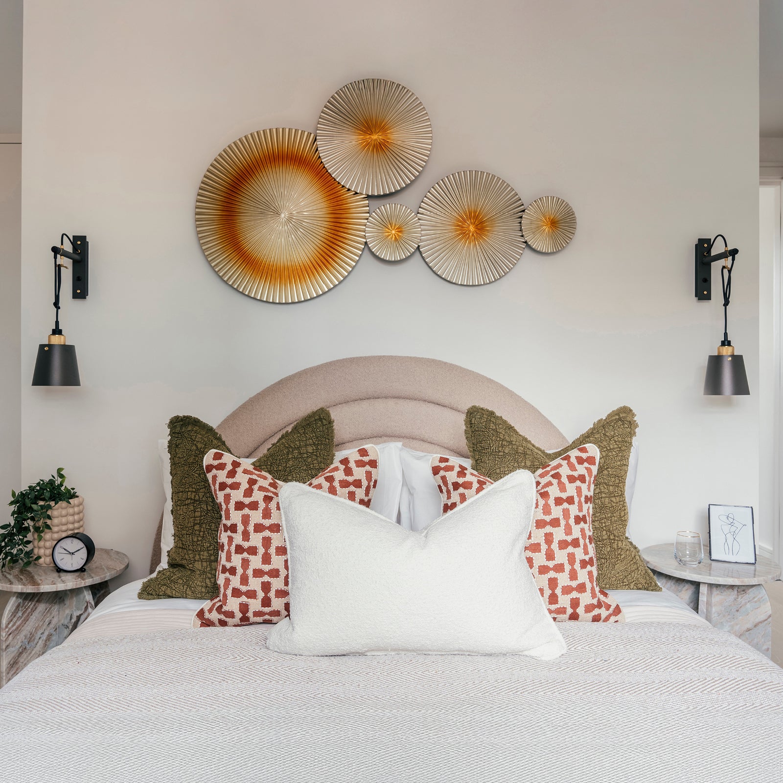 Stylish bedroom interior featuring a curved upholstered headboard, a neatly made bed with layered pillows in olive green, burnt orange, and white textures, and symmetrical round marble-top nightstands. Black wall-mounted reading lamps flank both sides of the bed, and a striking metallic wall sculpture with circular fan-like designs in gold and orange tones serves as a bold focal point above the bed.