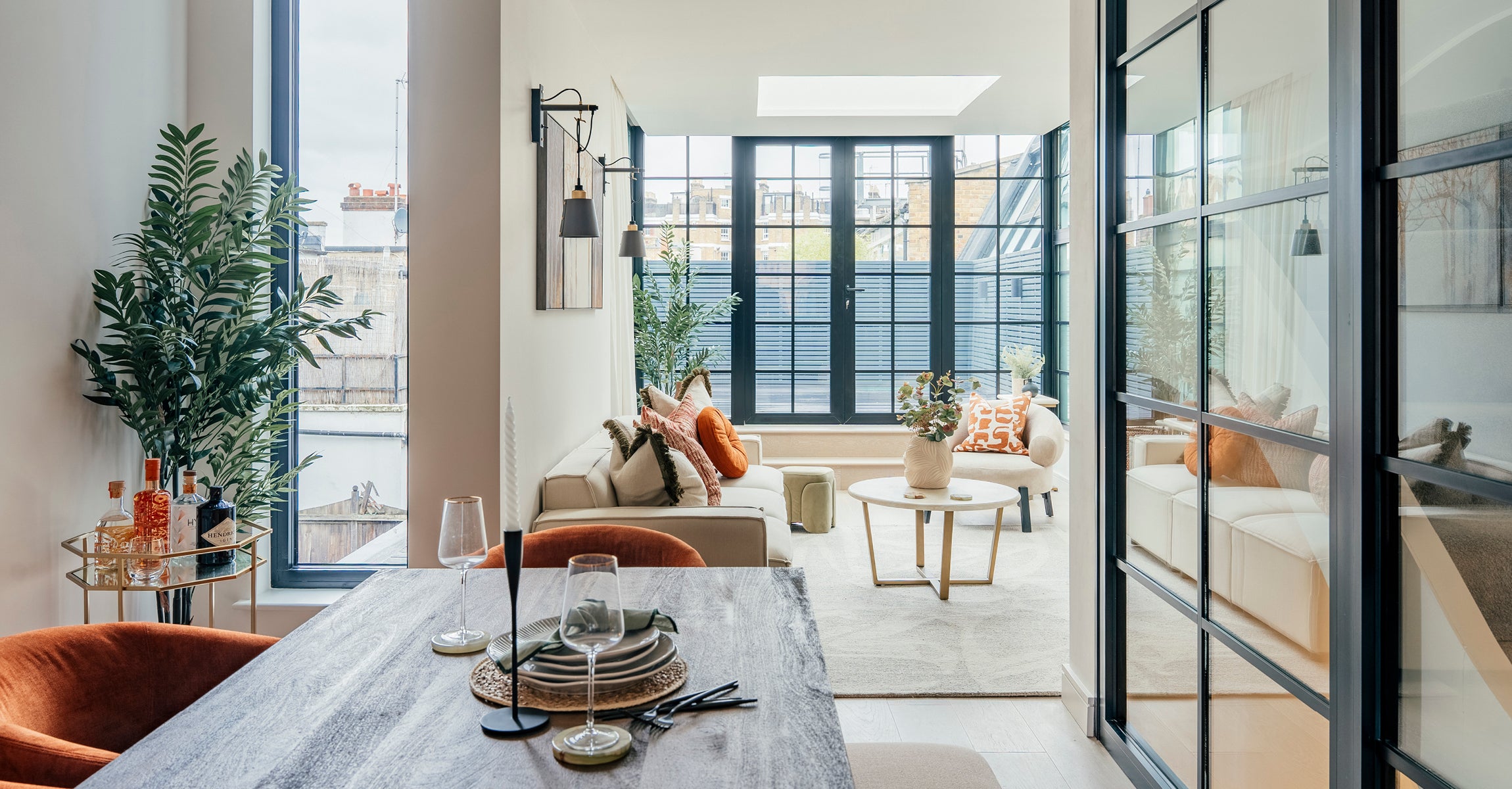 Stylish open-plan living and dining area featuring a cream sofa with orange and neutral cushions, a rustic wooden dining table set for two, large windows letting in natural light, and green plants adding a fresh, modern touch.