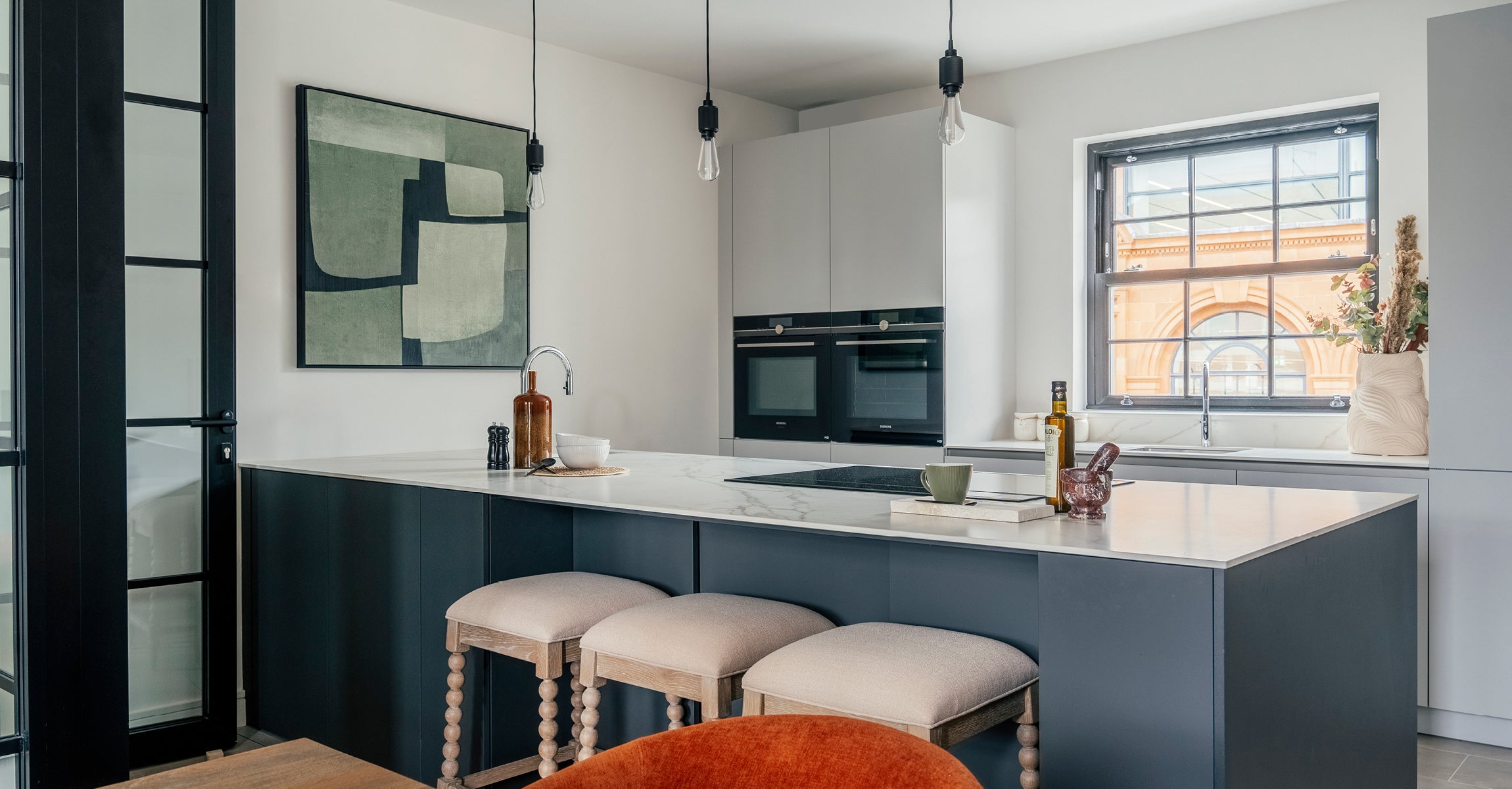 Modern kitchen with a large marble island and barstool seating, featuring sleek grey cabinetry, built-in ovens, and a window offering a view of a historic brick building, all accented by minimalist pendant lighting and contemporary decor.