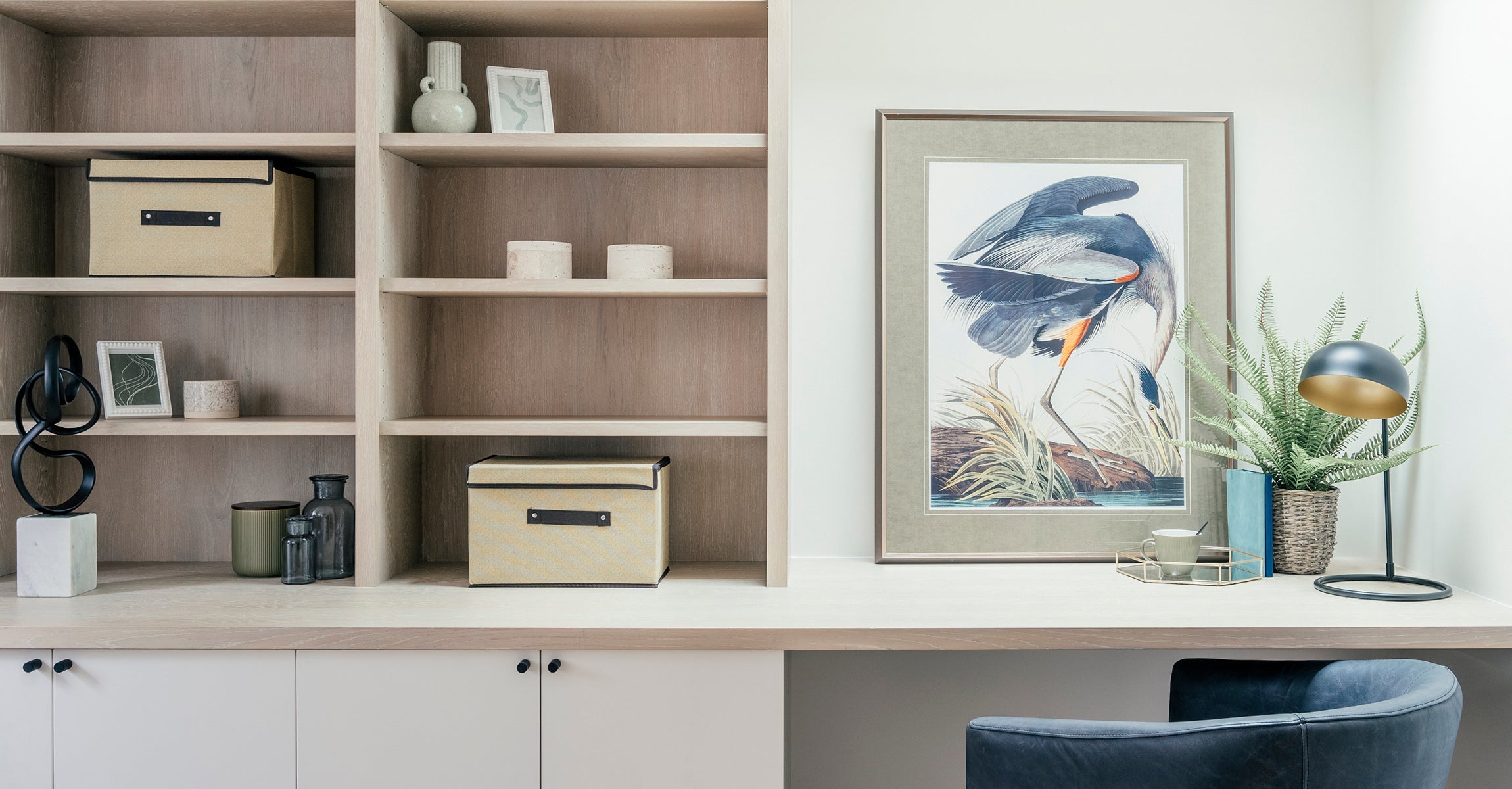 Stylish home office nook with built-in wooden shelving, decorative storage boxes, and a framed bird illustration, complemented by a modern desk lamp, potted plant, and a dark upholstered chair.