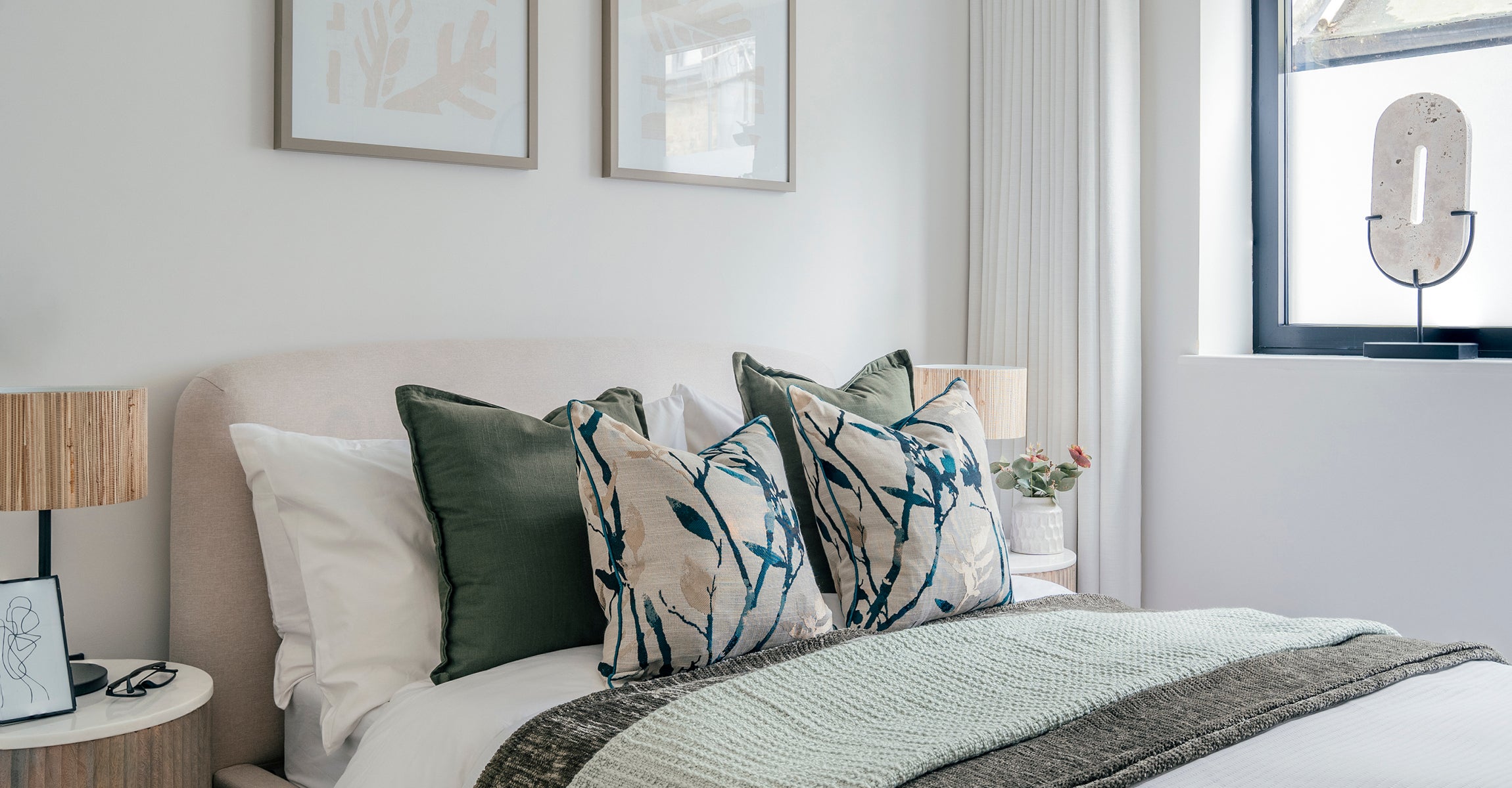 Bright and serene bedroom with a neutral upholstered headboard, layered green and botanical cushions, minimalist side tables, and soft natural light from a large window.