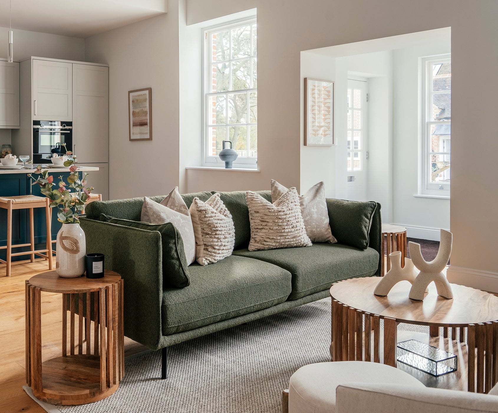 Modern open-plan living space with a green upholstered sofa adorned with textured neutral cushions, flanked by wooden slatted side tables. The room features light wooden flooring, a round wooden coffee table with a sculptural decor piece, and a view into a sleek kitchen with white cabinets and blue island. Large windows fill the space with natural light.