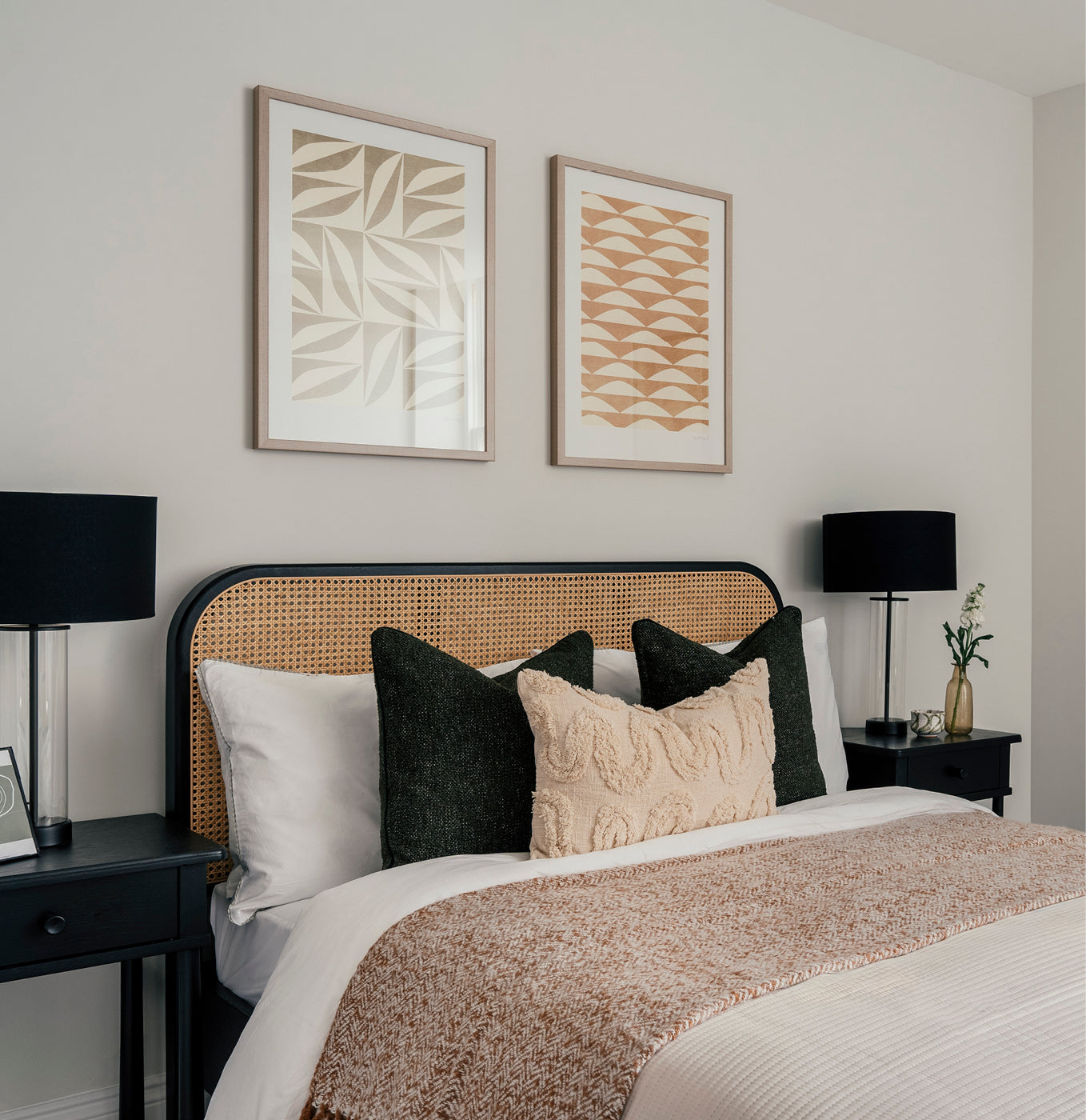 Stylish bedroom featuring a woven rattan headboard, neatly made bed with textured beige and green cushions, and a brown herringbone throw blanket. Two abstract art prints hang above the bed, and black bedside tables with matching black lamps and minimal decorative items frame the scene.