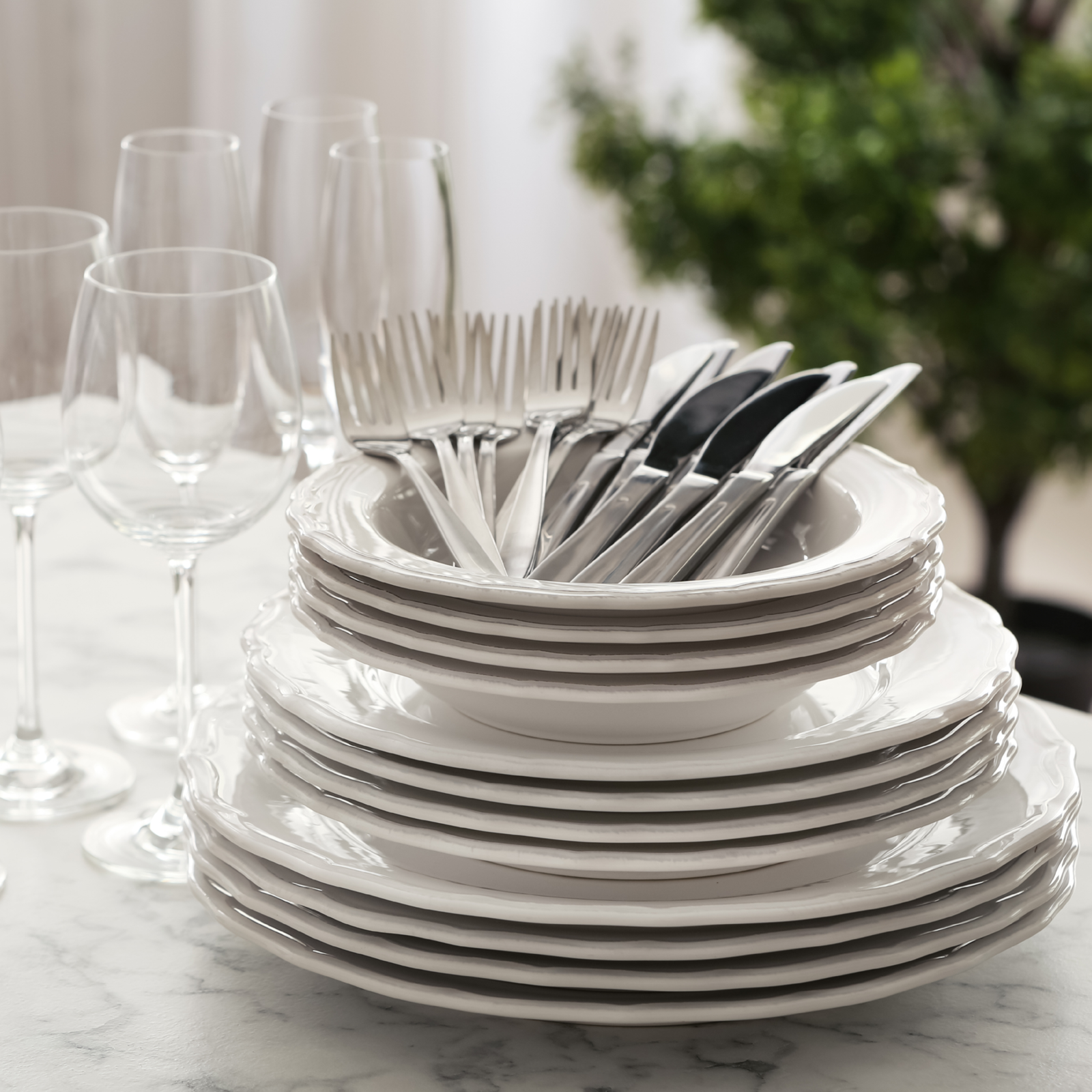Stack of white ceramic plates and bowls with stainless steel cutlery and wine glasses on marble countertop