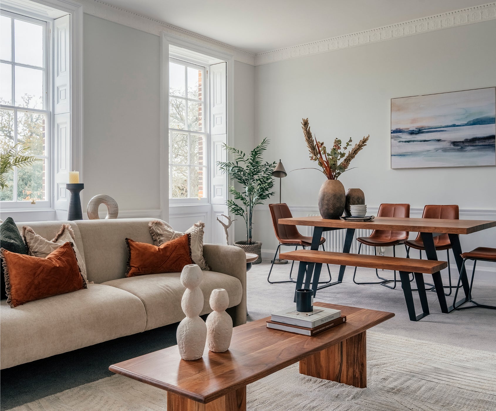 A bright and elegant open-plan living and dining room featuring a beige sofa with rust-coloured cushions, a wooden coffee table with stacked books and ceramic sculptures, and a dining area with a wooden table, matching bench, and leather chairs. Large sash windows let in natural light, while decorative vases, wall art, and indoor plants enhance the cozy and sophisticated aesthetic.