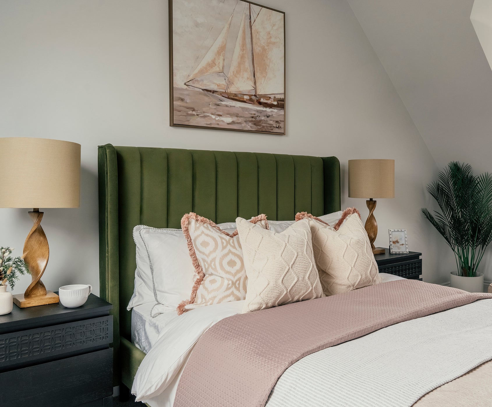 A stylish bedroom featuring a green vertical-panel upholstered headboard, layered with cream and patterned decorative pillows and a soft blush pink bedspread. Matching black nightstands with carved detailing flank the bed, each topped with a twisted wooden lamp with beige shades. Above the bed hangs a nautical-themed sailboat painting, and a potted palm adds a natural touch in the corner.
