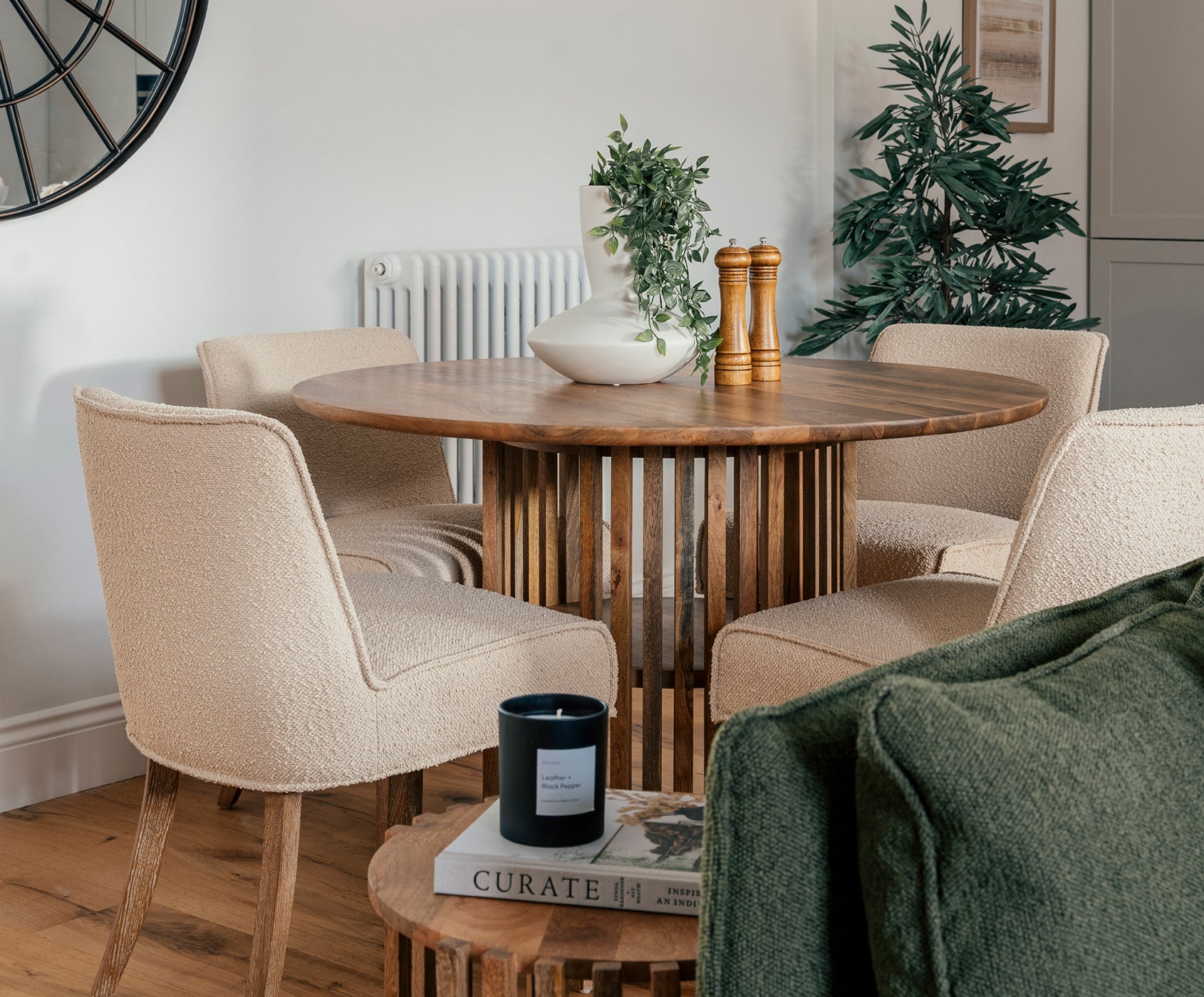 Cozy dining area featuring a round wooden table with slatted pedestal base, surrounded by four textured cream upholstered chairs, complemented by greenery and warm decorative accents.