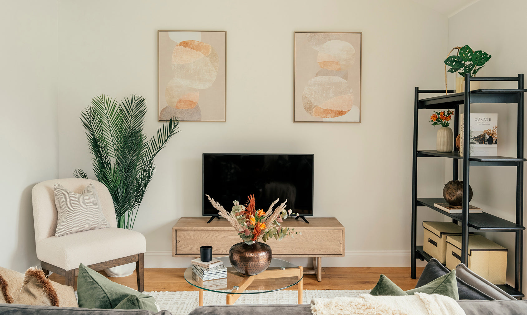 Stylish living room with neutral tones, abstract wall art, modern wooden media unit, decorative shelving, and a glass coffee table with floral arrangement.