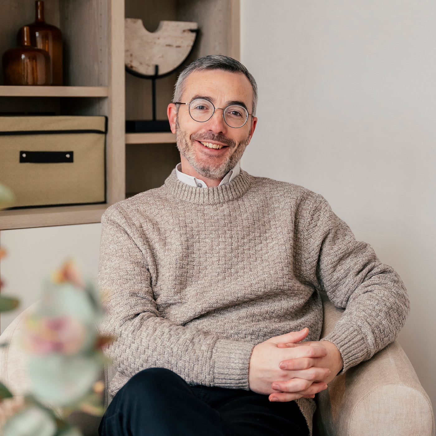 Smiling man with short gray hair and glasses wearing a beige sweater seated in a modern office with minimalist shelving