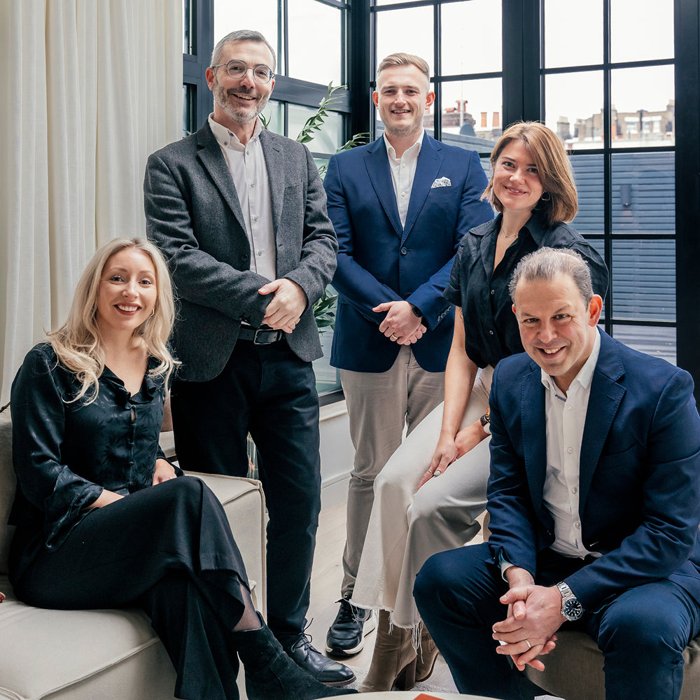 Professional group photo of five team members in stylish business attire, posing in a modern office with large windows and natural light