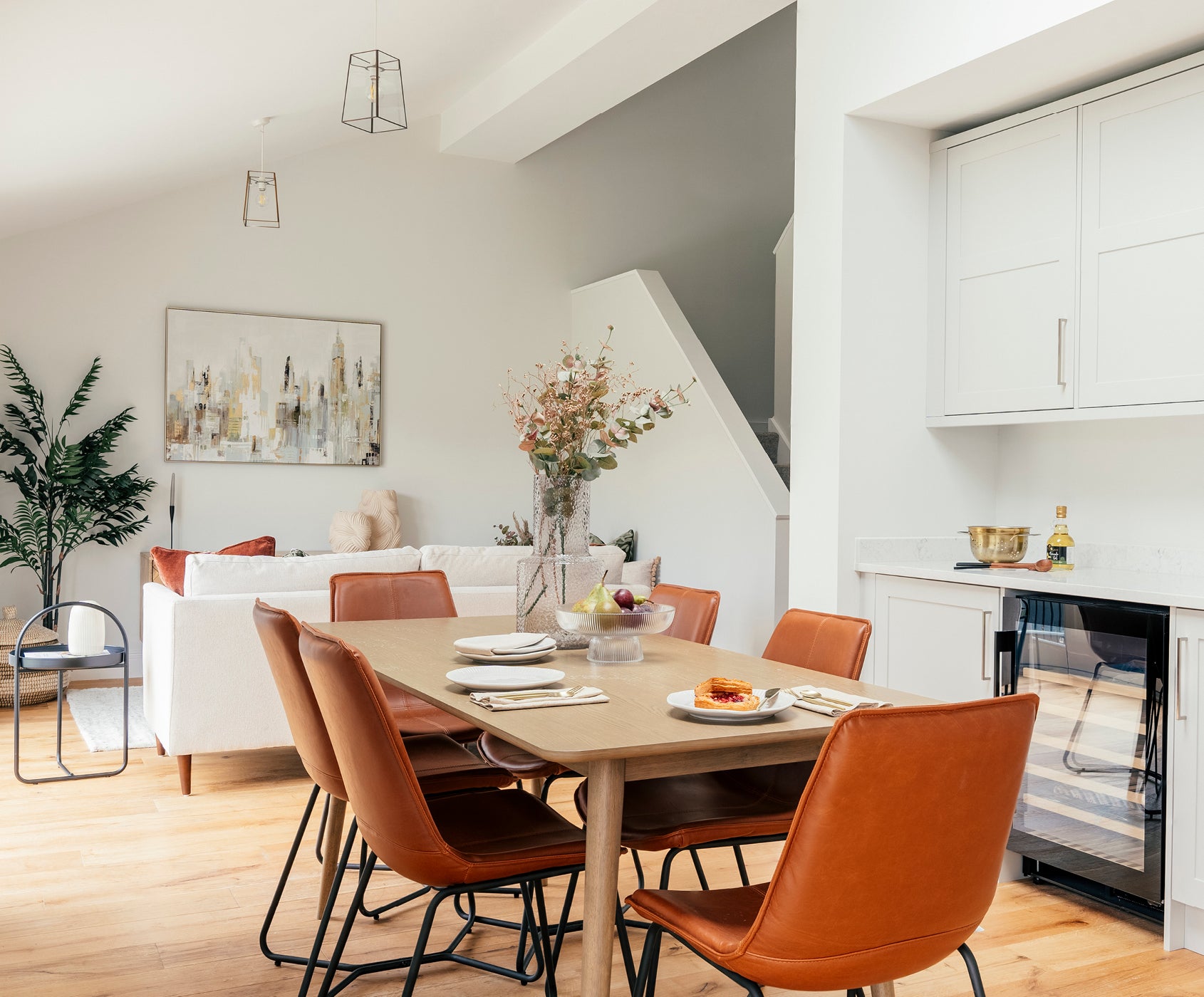 Modern open-plan dining and living area featuring a wooden dining table surrounded by six burnt-orange leather chairs, minimalist white cabinetry with a built-in wine fridge, a white sofa in the background, and contemporary decor including abstract artwork and a tall potted plant.