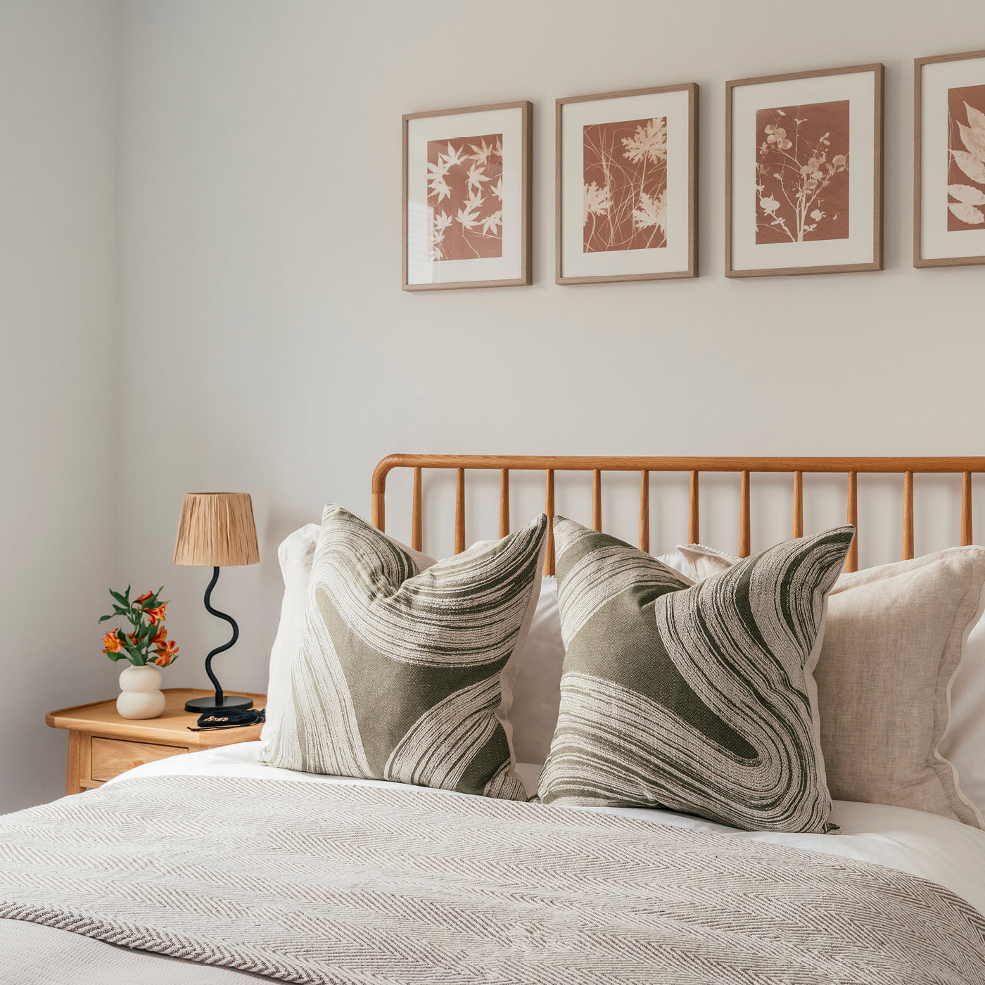 Cozy bedroom with a wooden slatted headboard, two large abstract-patterned green pillows, neutral bedding, and a minimalist nightstand featuring a sculptural lamp and small floral arrangement; above the bed hang four botanical art prints in warm tones.