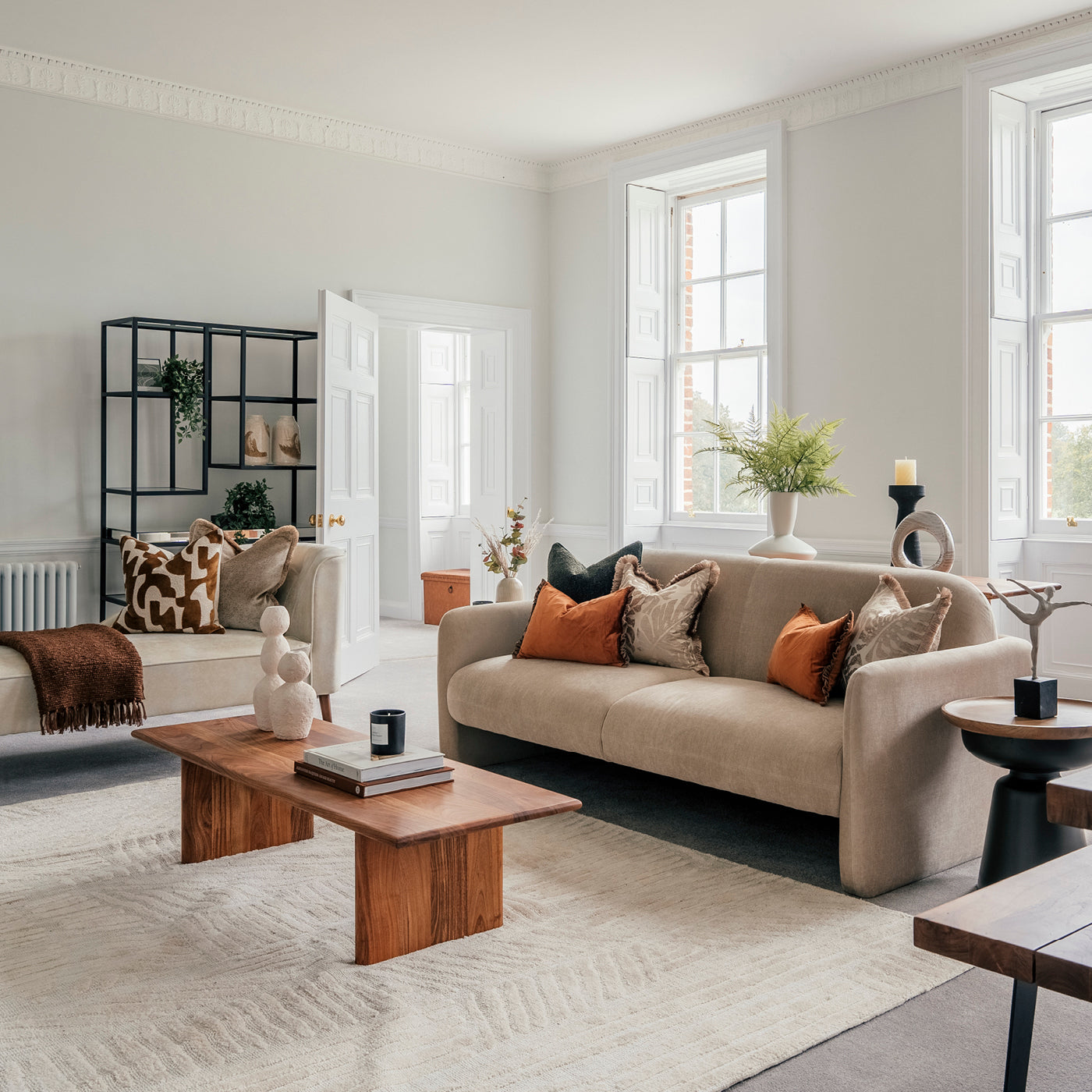 Elegant living room with large sash windows, neutral-toned furniture, and earthy accent pillows; features a beige sofa, wooden coffee table, sculptural decor, and a modern black shelving unit with curated ornaments.