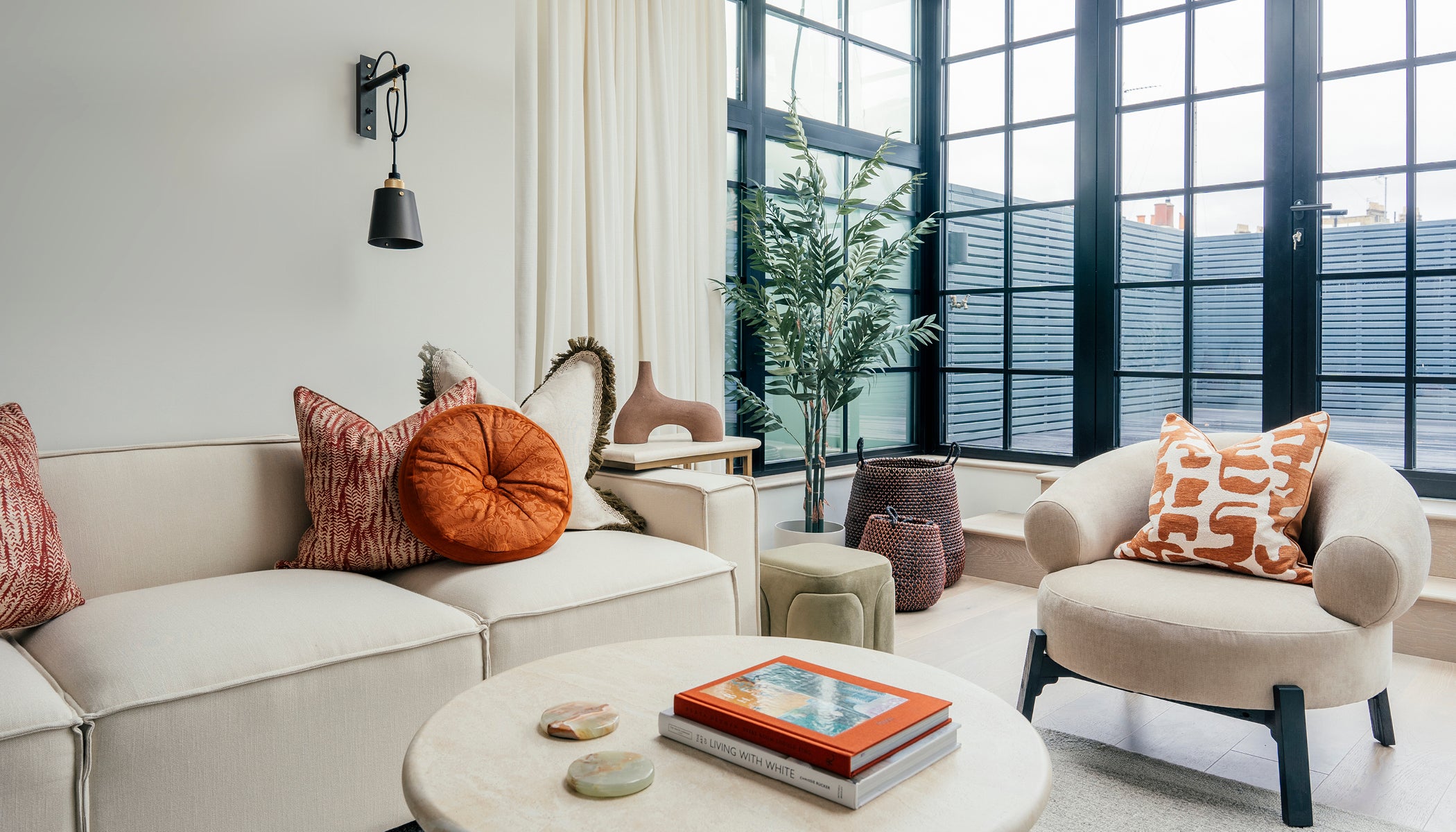 Bright living room with cream furniture, colourful cushions, large grid-style windows, and modern decor elements like stacked books and indoor plants.