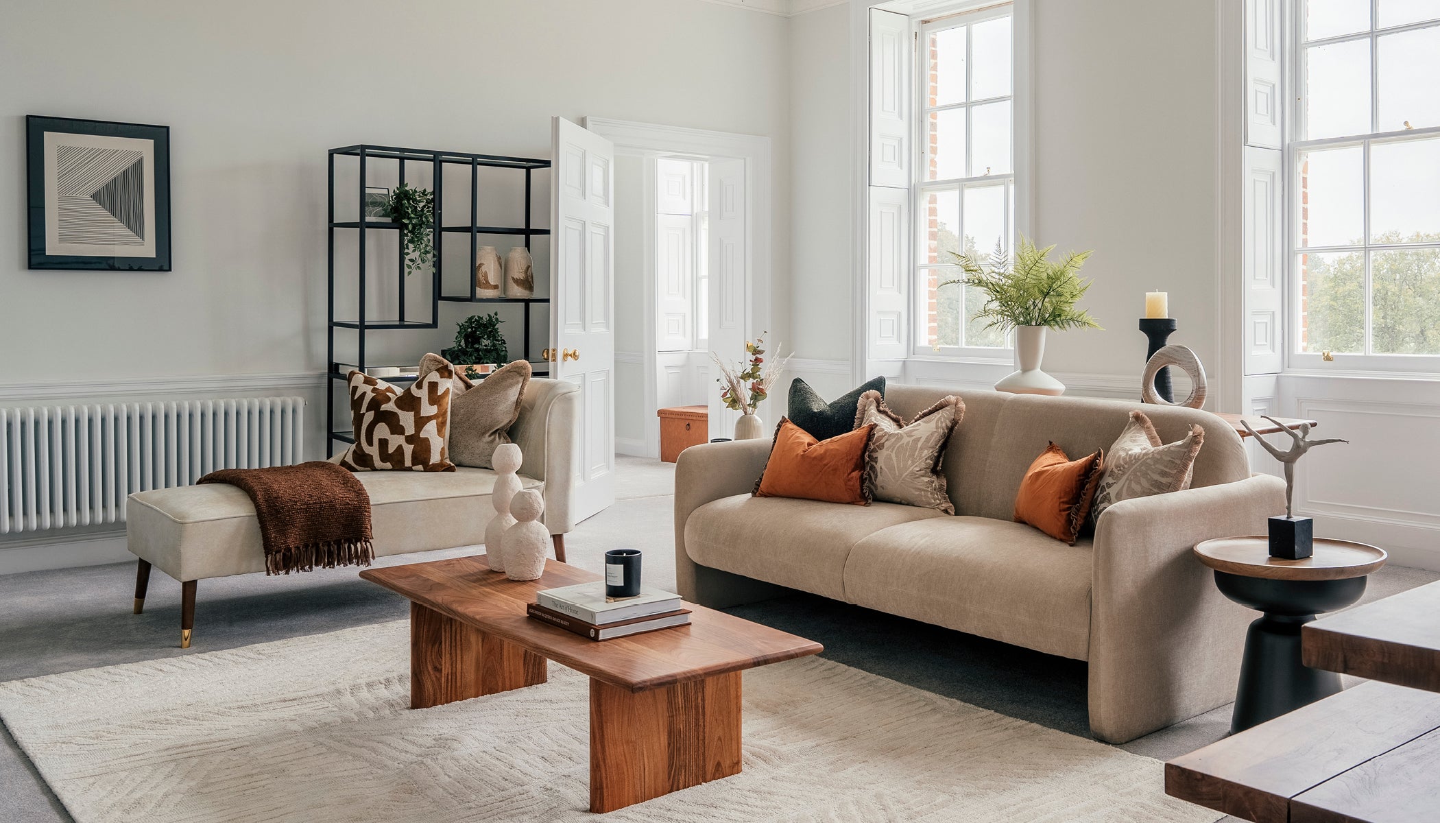Spacious and elegantly decorated living room featuring neutral-toned sofas with warm accent pillows, a wooden coffee table with sculptural decor, large sash windows letting in natural light, and modern shelving with minimalist accessories.