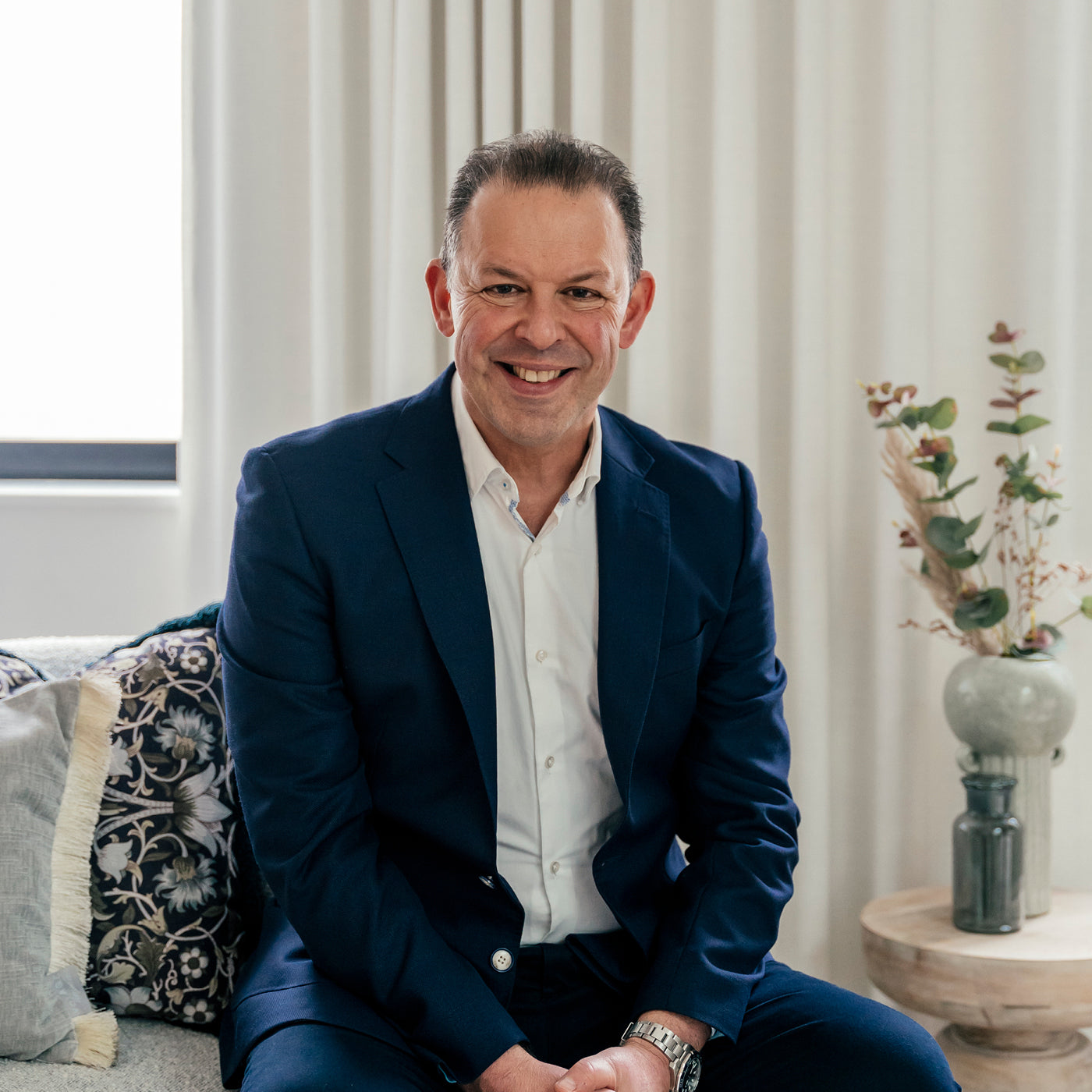 Confident man in a navy blue suit smiling while seated in a modern, light-filled office with neutral decor